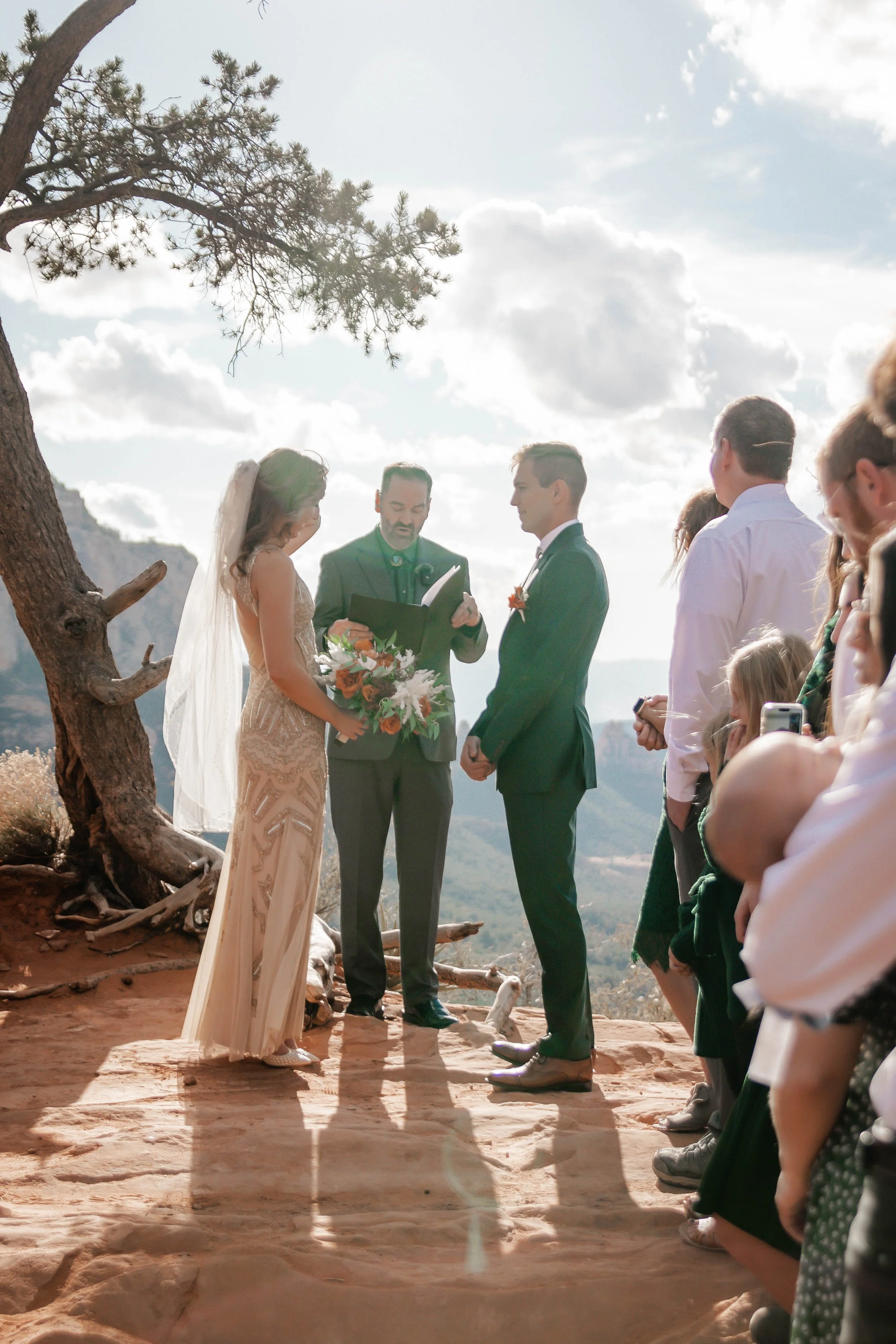 A wedding ceremony outdoors on a rocky terrain with mountains and cloudy sky in the background. The bride and groom stand before an officiant, holding hands. The bride is in a lace dress with a veil, holding a bouquet. The groom wears a dark suit. Gu
