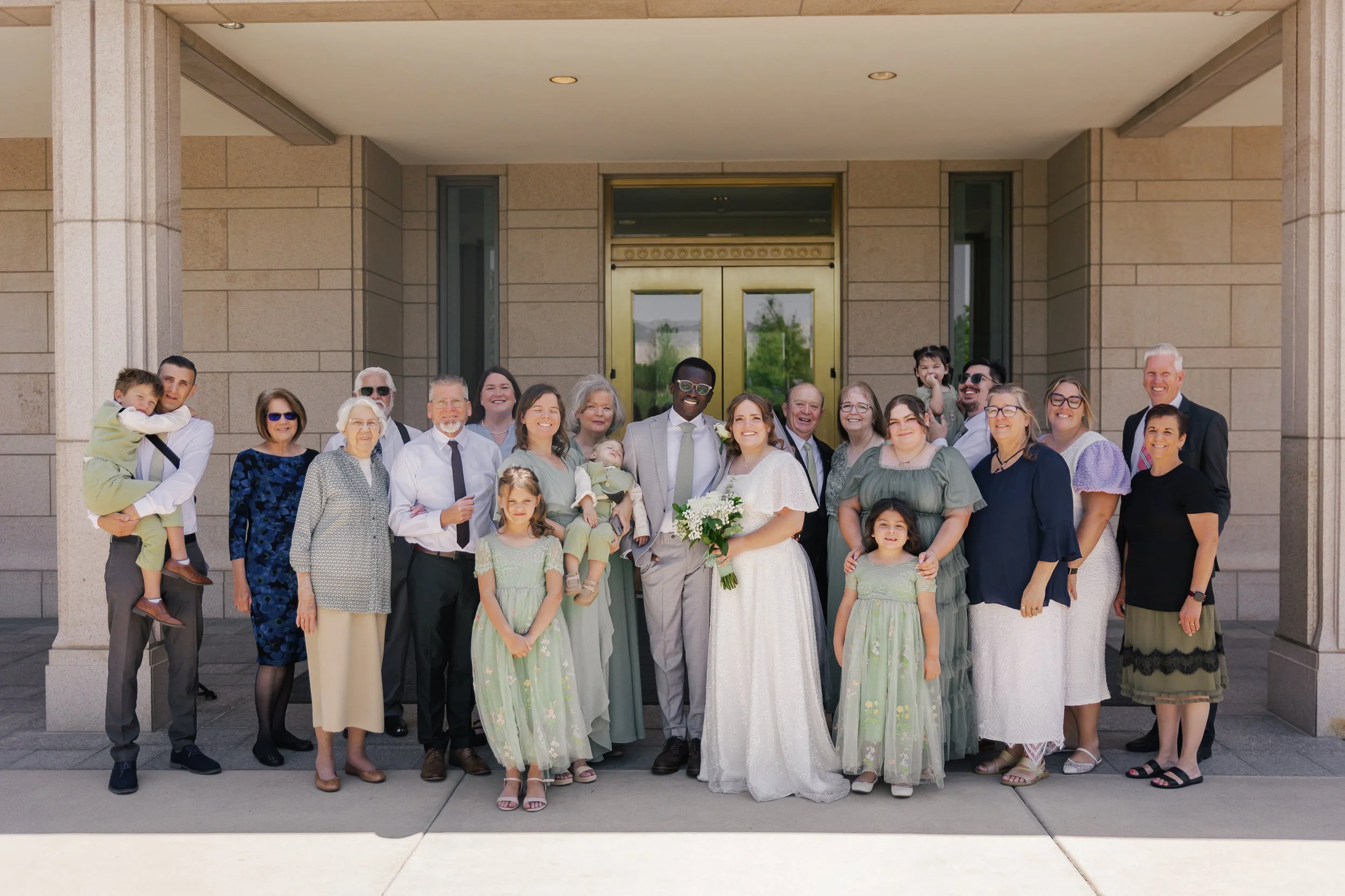 A large group of people gathered outside a building, celebrating a wedding. The bride and groom are at the center, smiling and holding a bouquet of flowers. The wedding party and family are surrounding them, dressed in formal and semi-formal attire, 