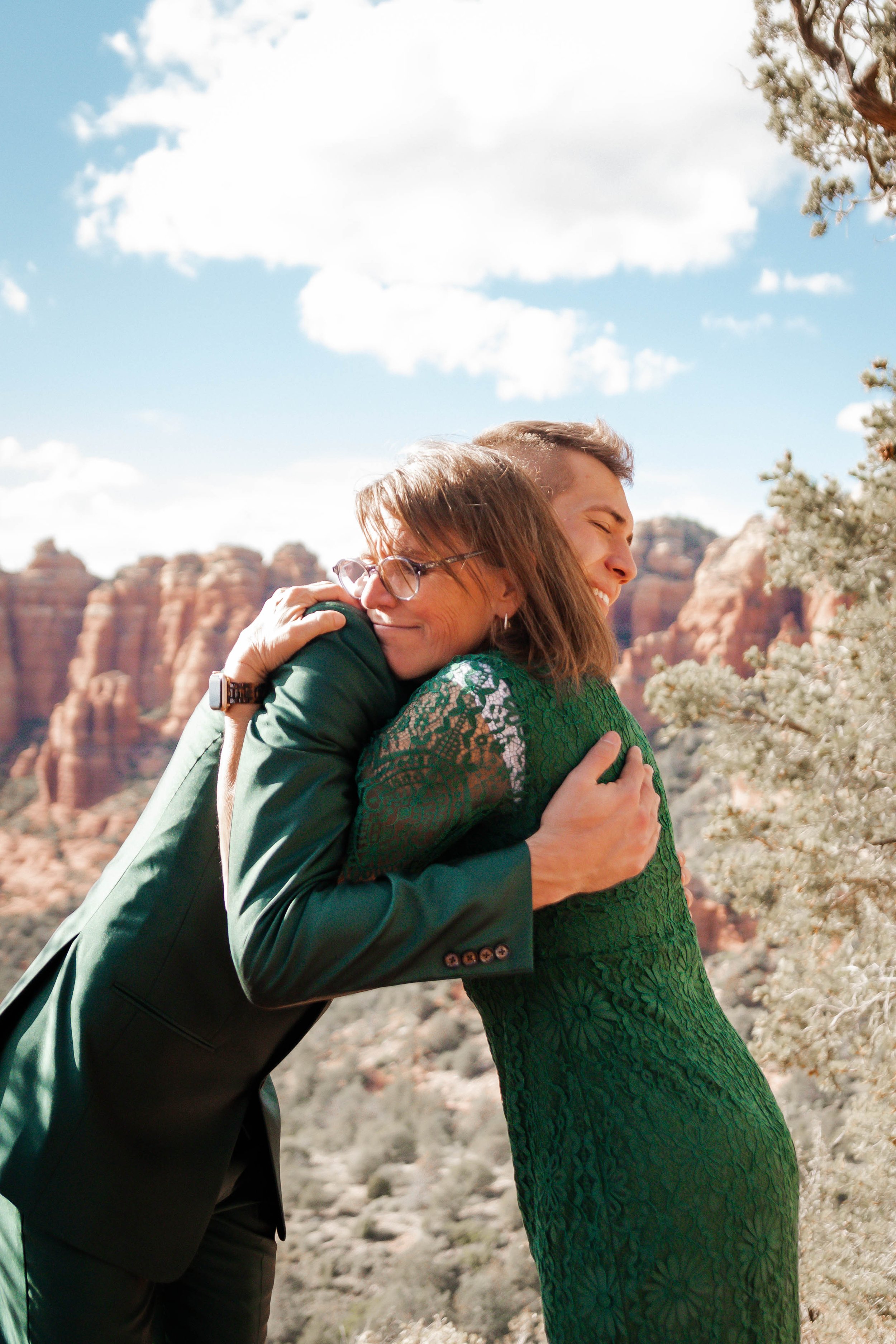 Two women sharing an embrace outdoors on a sunny day, with a mountain desert landscape and blue sky in the background.