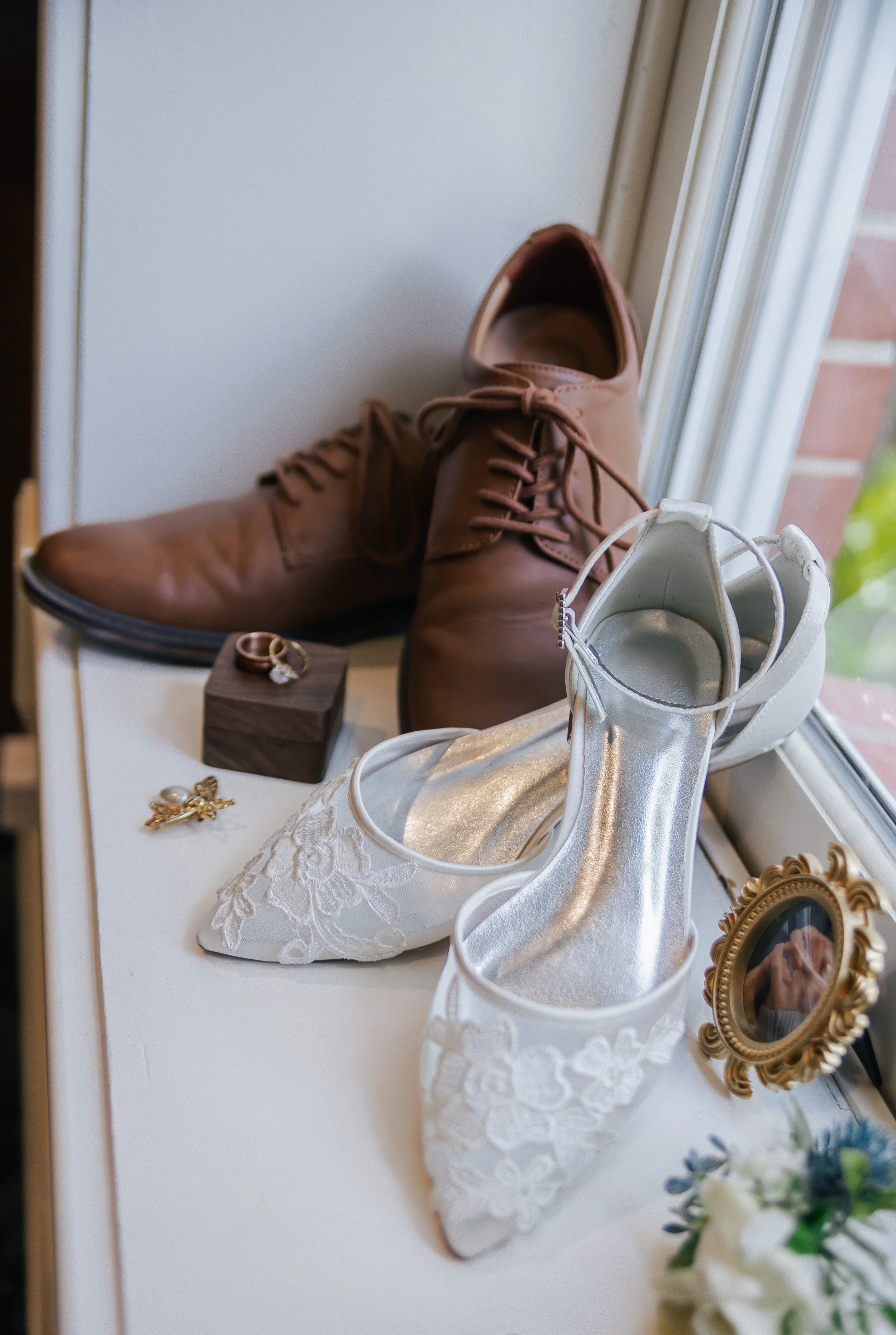 Brown dress shoes, white embroidered wedding shoes, wedding rings, a gold wedding band, a small picture frame, and a floral arrangement on a white surface near a window.