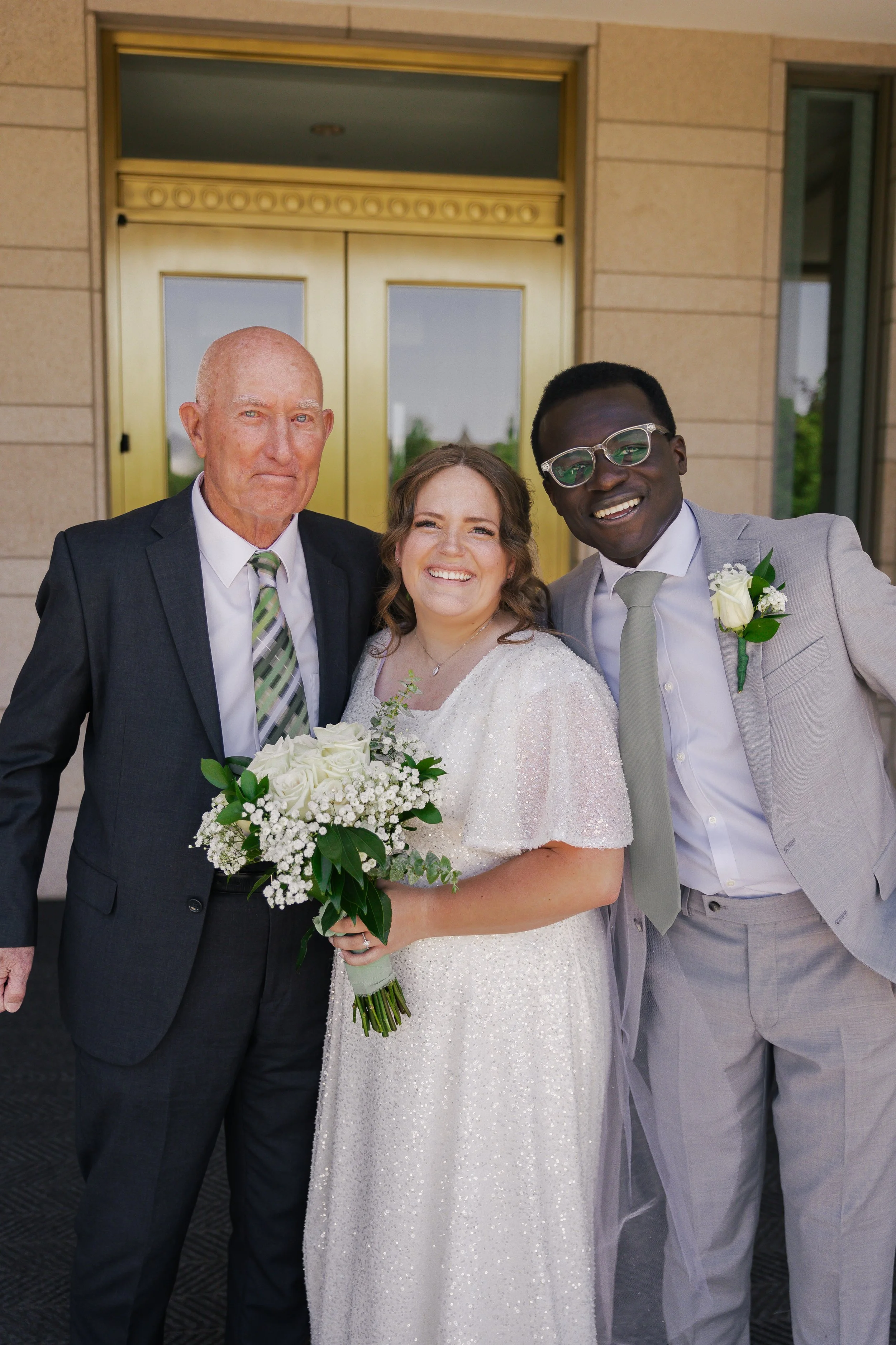 Three people dressed in wedding attire standing outside a building, smiling at the camera. The woman in the center is holding a bouquet of white flowers. The man on the left is wearing a dark suit and patterned tie. The man on the right is wearing a 