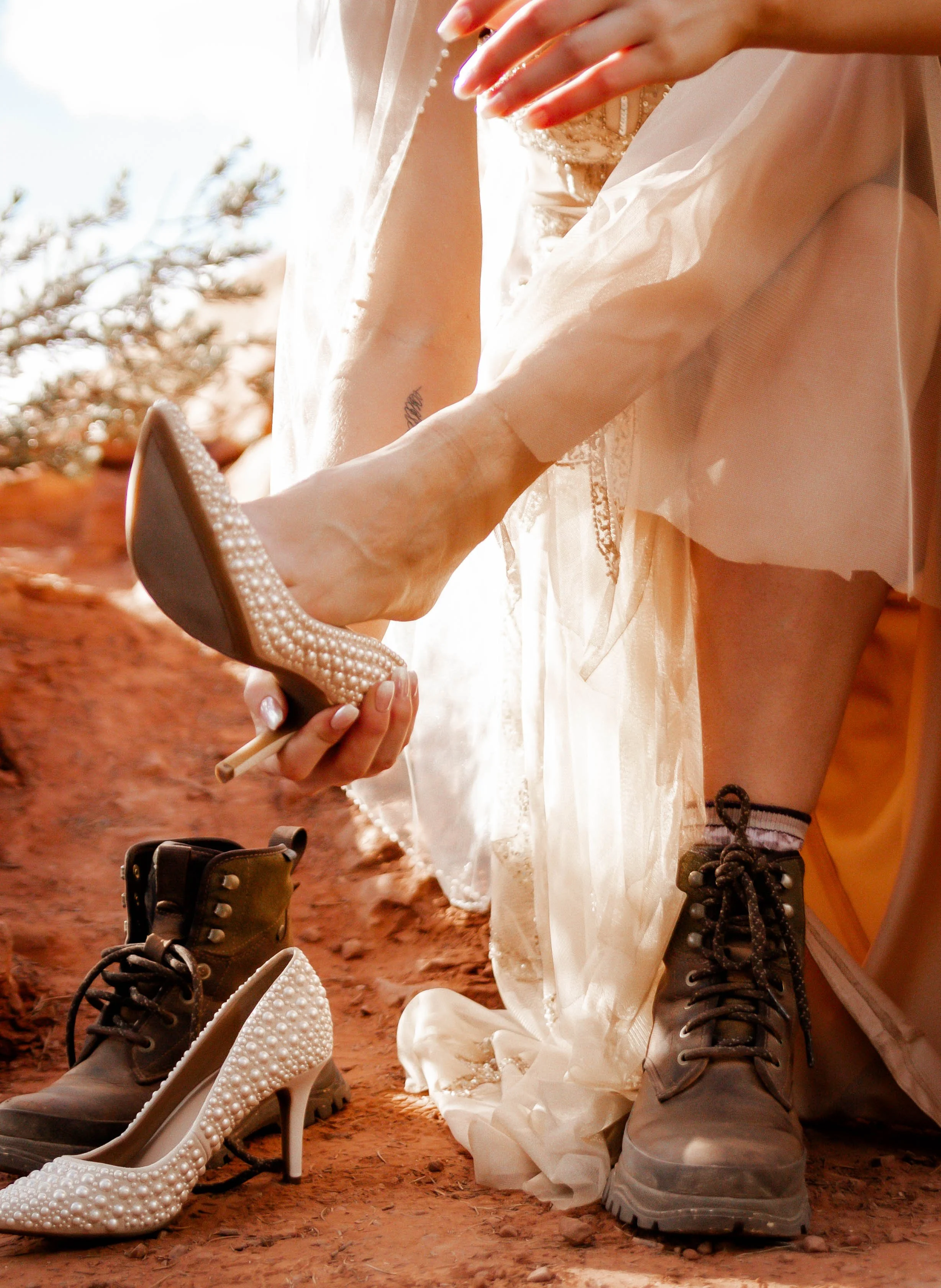 A person in a wedding dress is trying on a pearl-embellished high-heeled shoe while sitting on the ground in an outdoor setting with dirt and sparse vegetation.