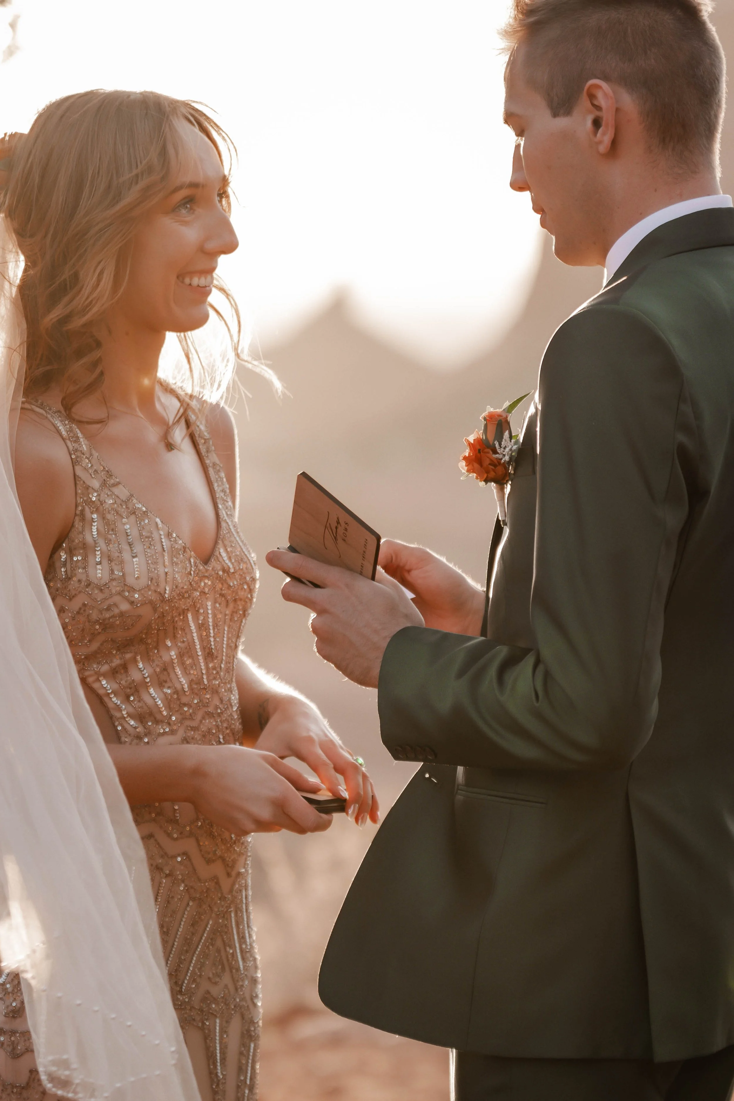 A bride and groom exchanging vows outdoors during sunset, with the groom reading from a card.