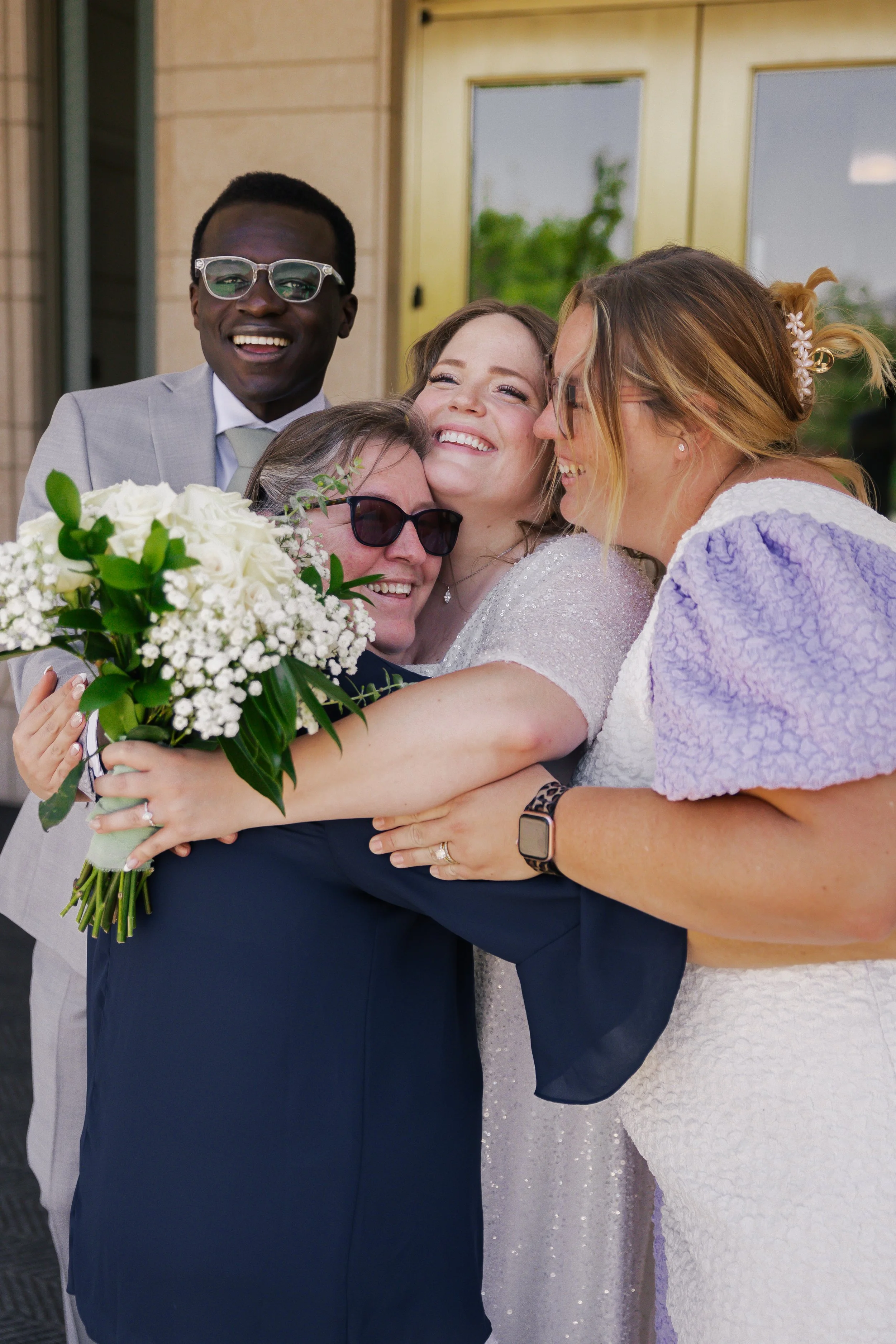 A group of five women hugging and smiling at a celebration, with one woman holding a bouquet of white flowers, standing outside a building with glass doors.