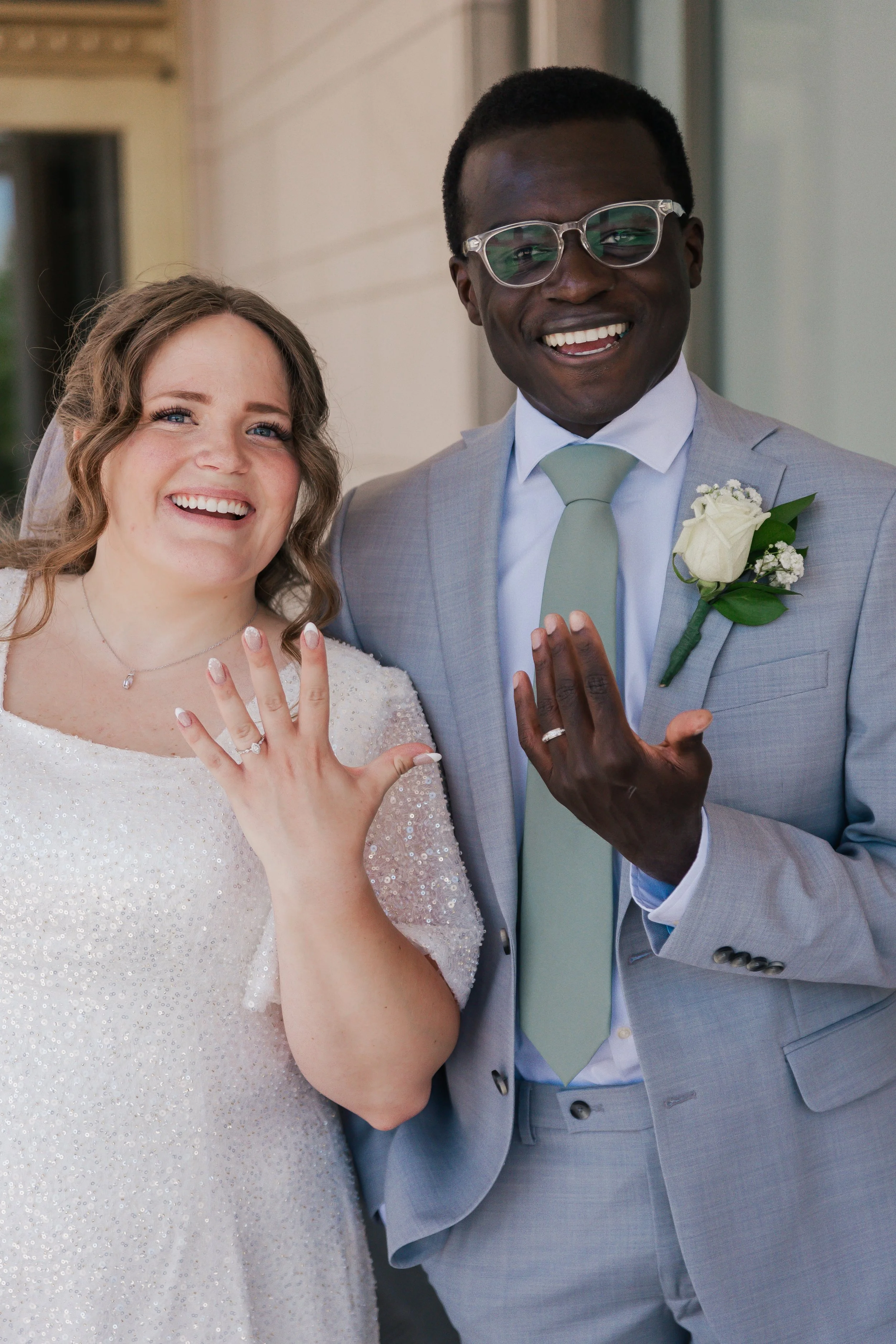 A bride and groom smiling and displaying their wedding rings outside a LDS temple.