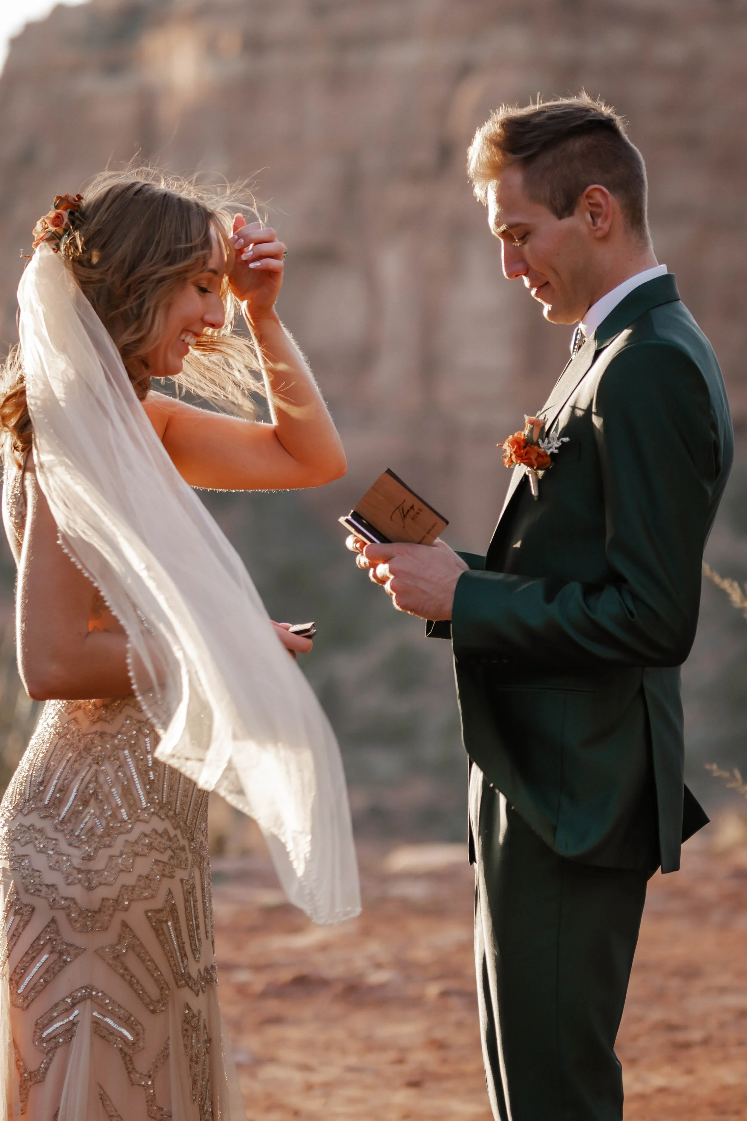 A bride and groom exchanging vows outdoors at sunset, with the groom reading vows from a booklet, and the bride smiling and touching her hair, both dressed in formal wedding attire.