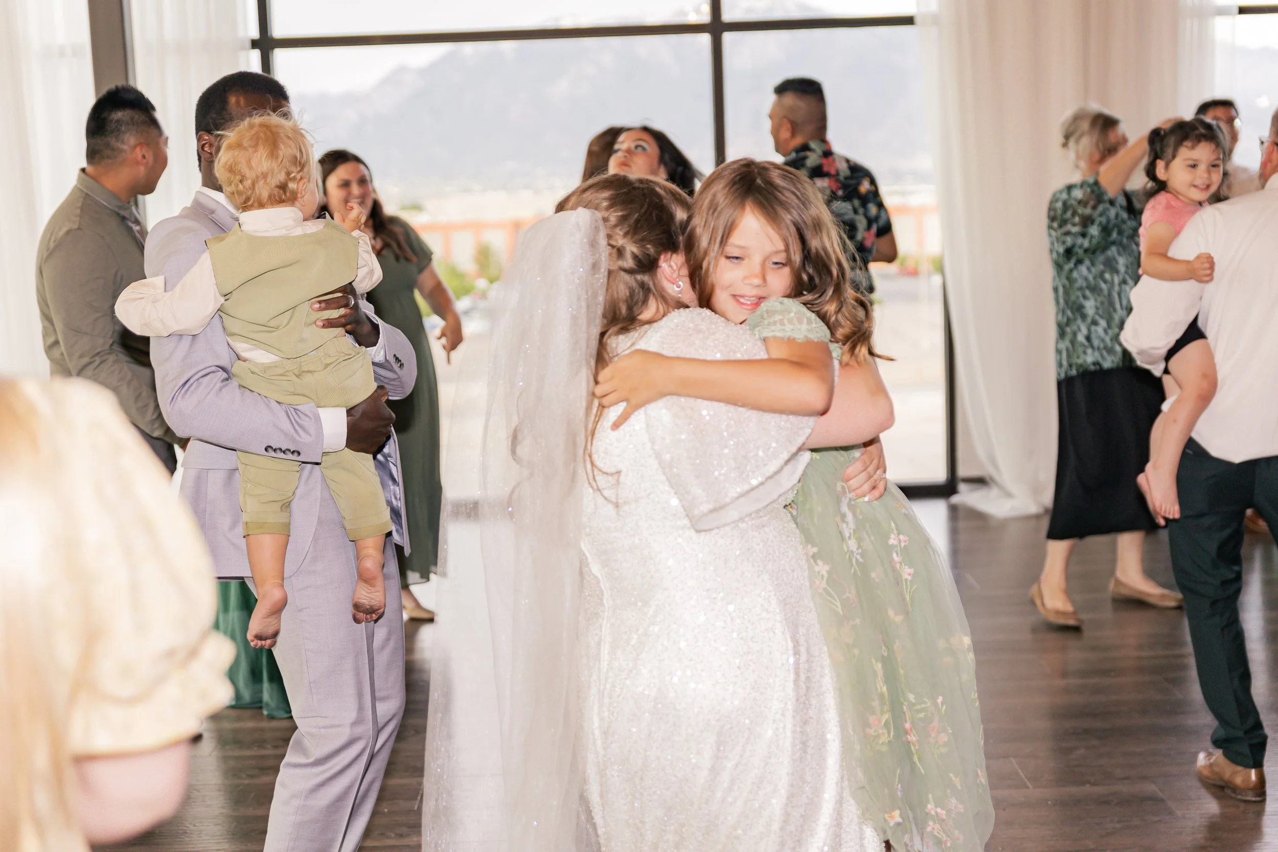 People dancing and hugging at a wedding reception indoors with large windows in the background.