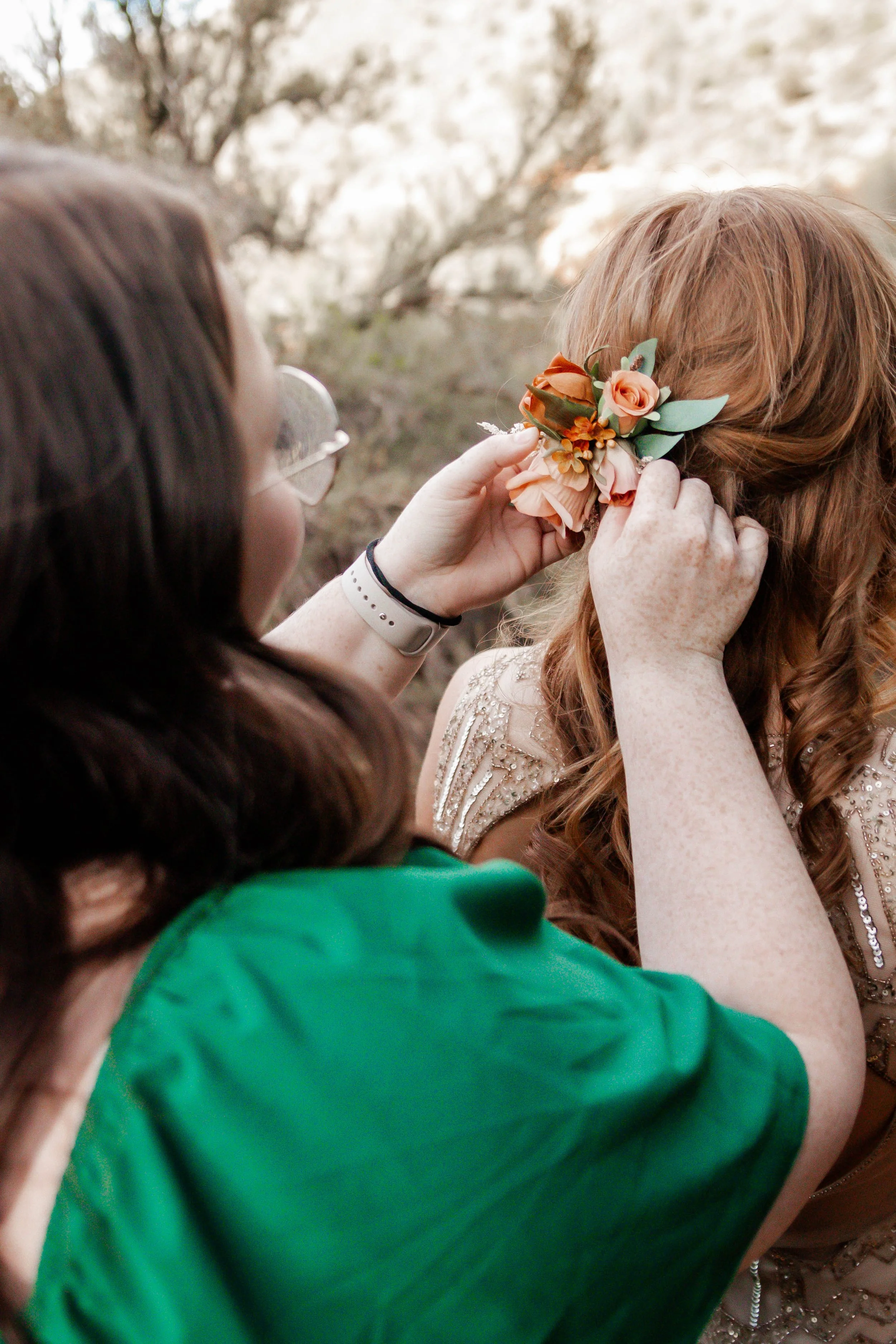 A woman with glasses and a white wristband places a floral hair accessory with peach-colored flowers and green leaves in another woman's long red hair, outdoors with rocky terrain in the background.