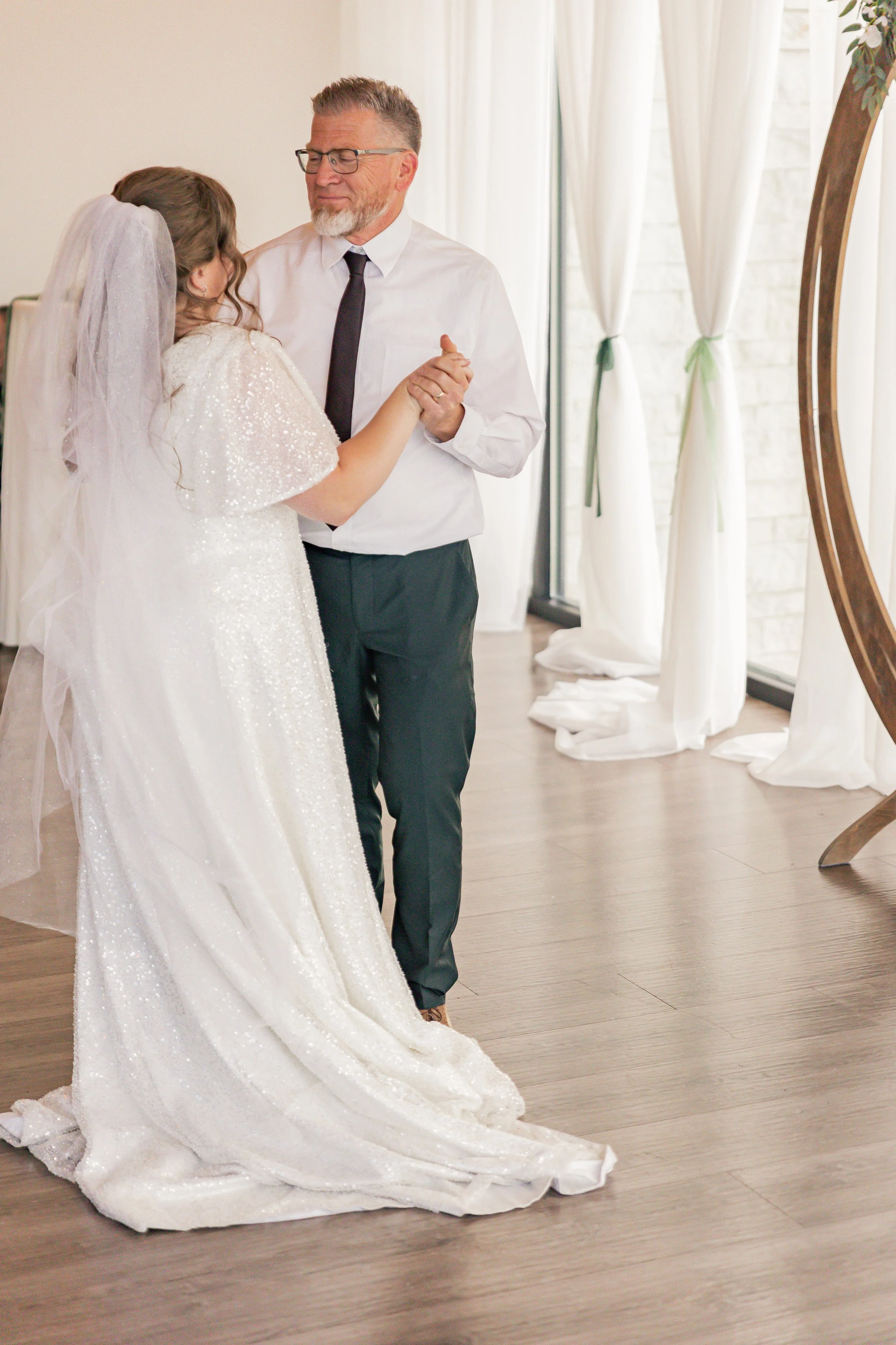 A bride dancing with an older man, possibly her father, during a wedding reception. She is wearing a white wedding gown with a veil, and they are holding hands while dancing indoors near a large window with white curtains.