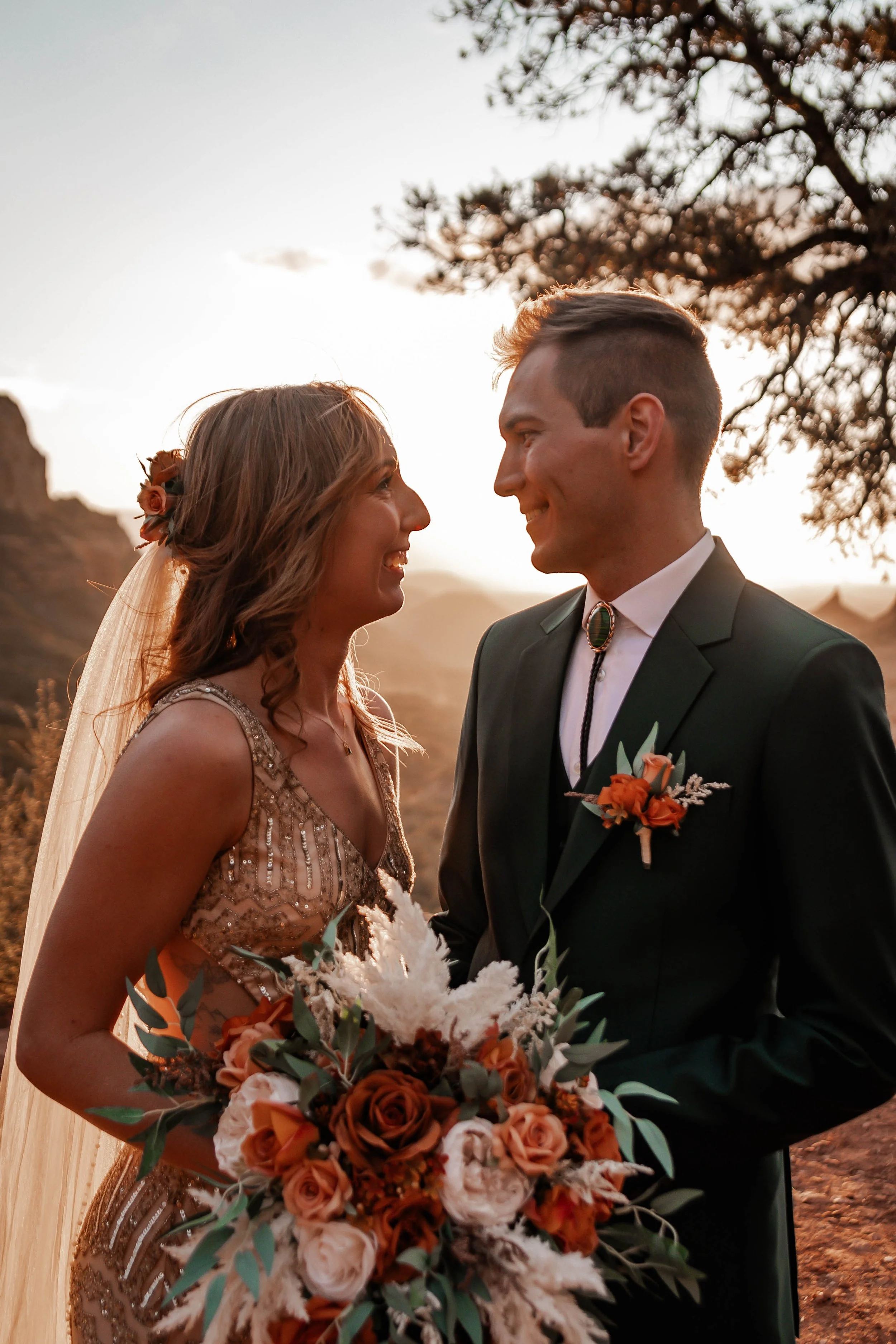 A bride and groom smiling at each other outdoors during sunset, with the bride holding a bouquet of flowers and the groom wearing a suit with a flower boutonniere.