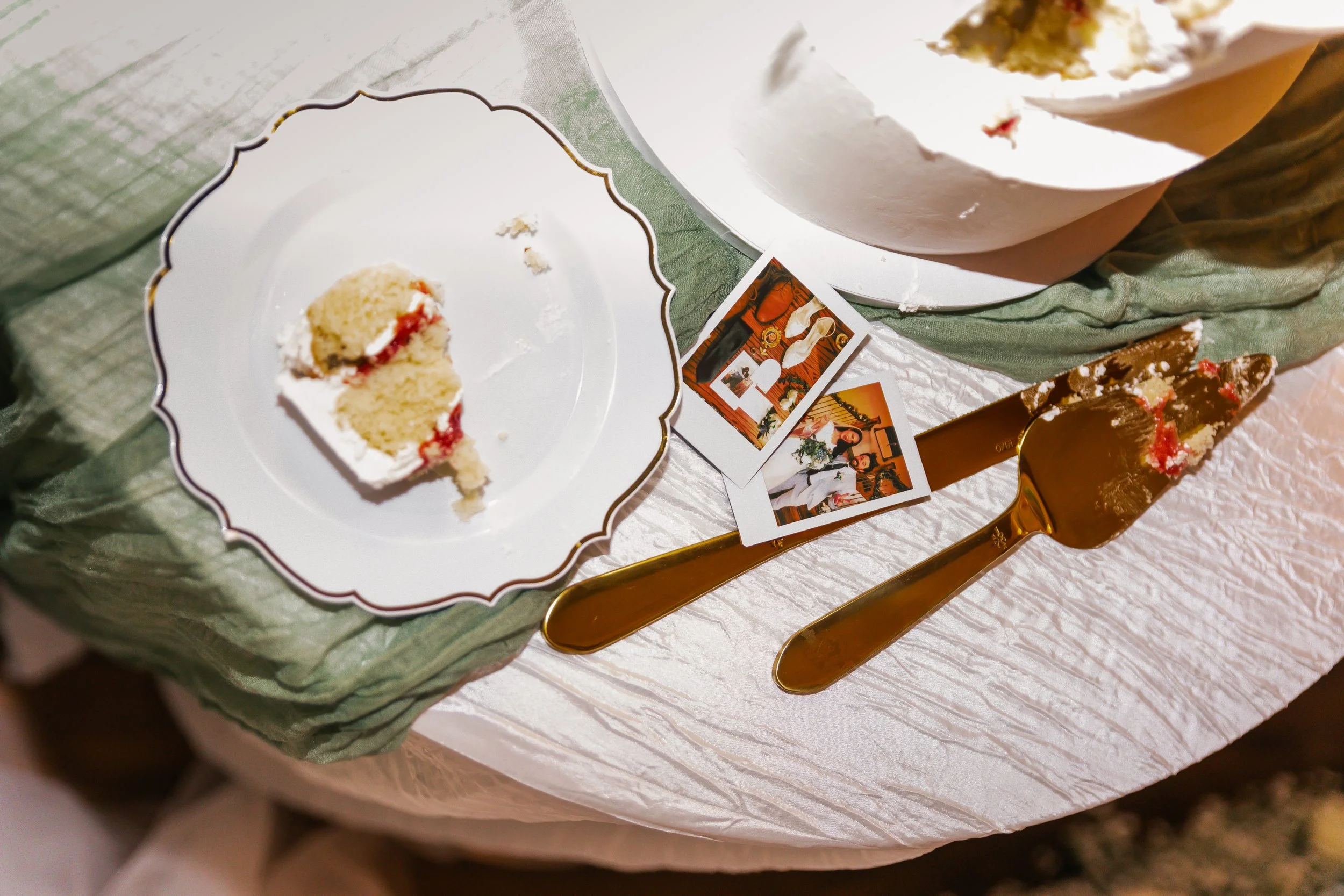 A partially eaten slice of cake with white frosting and red filling on a decorative white plate. The plate is on a table with a green cloth and two vintage-style photographs, a cake server, and a gold-colored knife and fork with pieces of cake on the