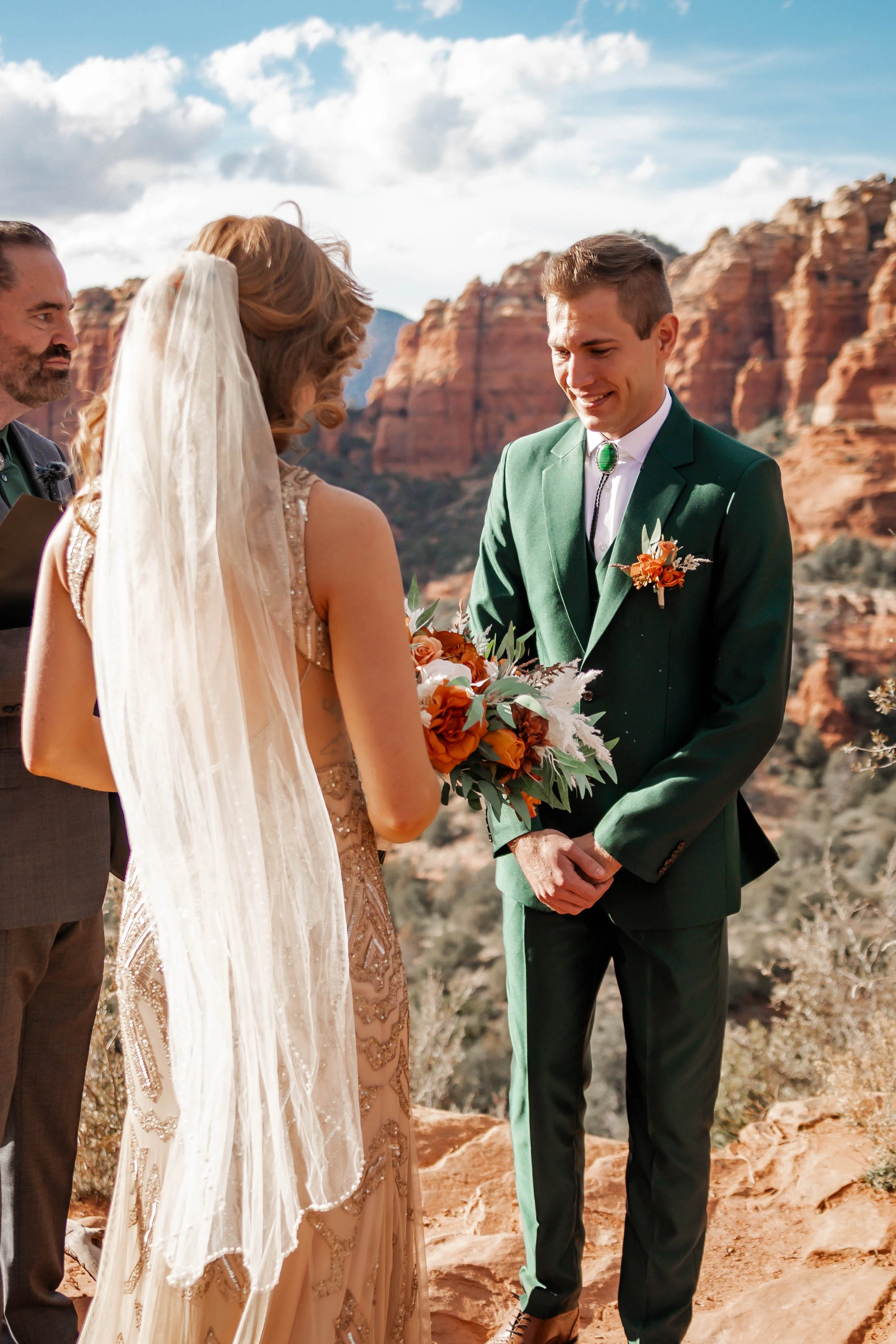 A couple getting married outdoors at a scenic desert location with red rock formations, during daytime with a partly cloudy sky.