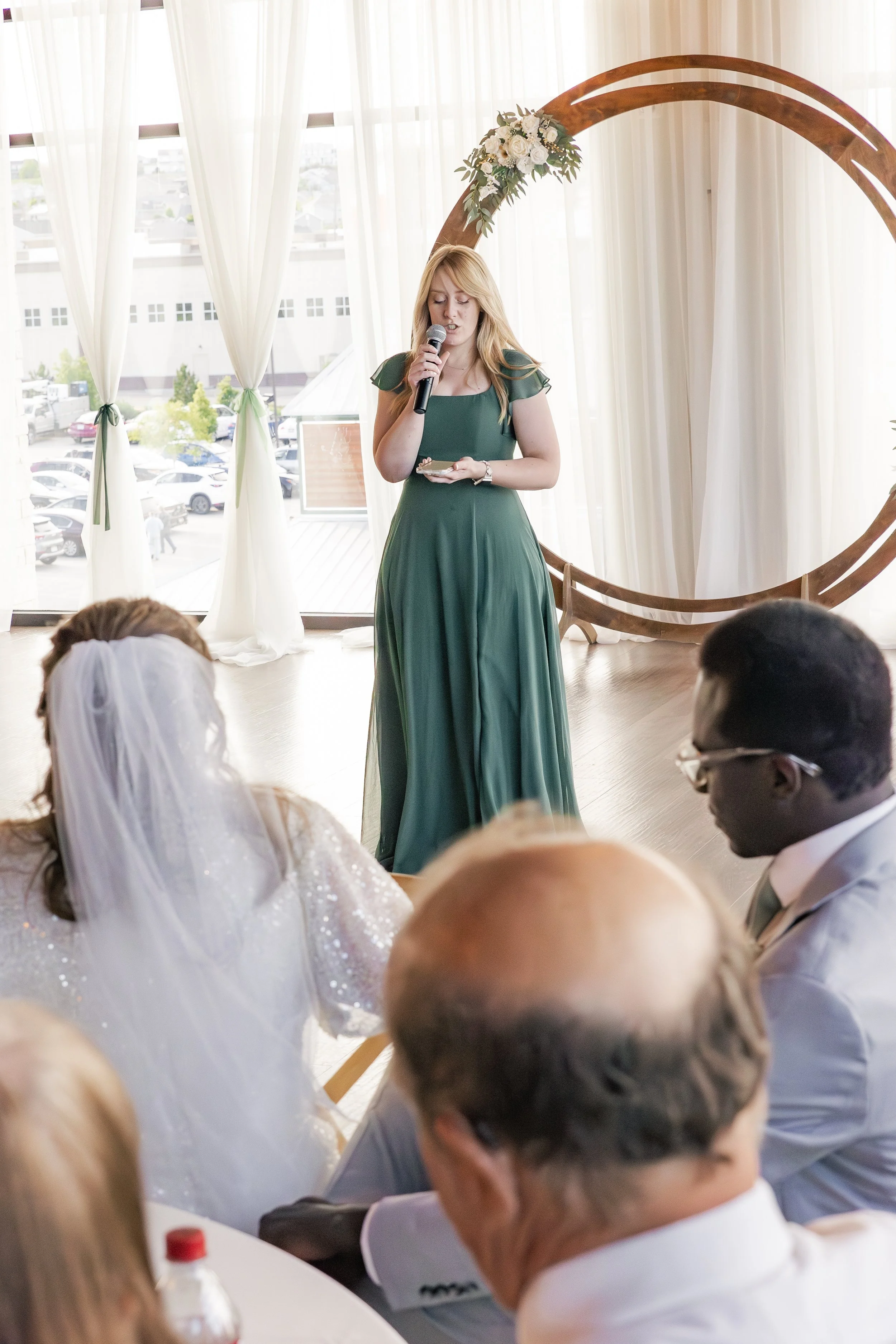 A woman in a long green dress giving a speech at a wedding, with a microphone in hand, in front of a decorated circular wooden arch with flowers, and seated guests including the bride with a veil and the groom in glasses.