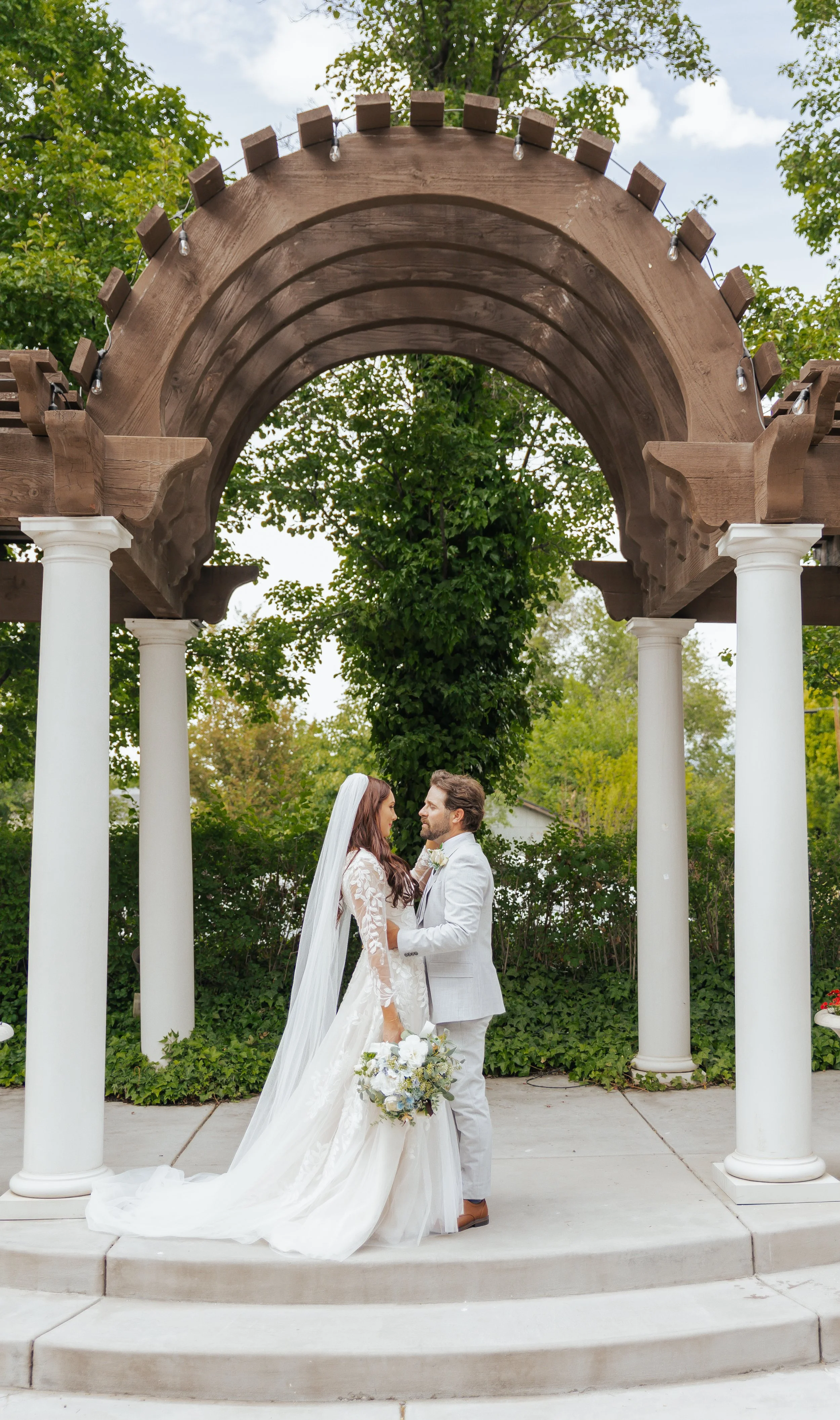 A bride and groom standing under a decorative wooden arch with white columns, on a wedding ceremony outdoors, with trees and greenery in the background.