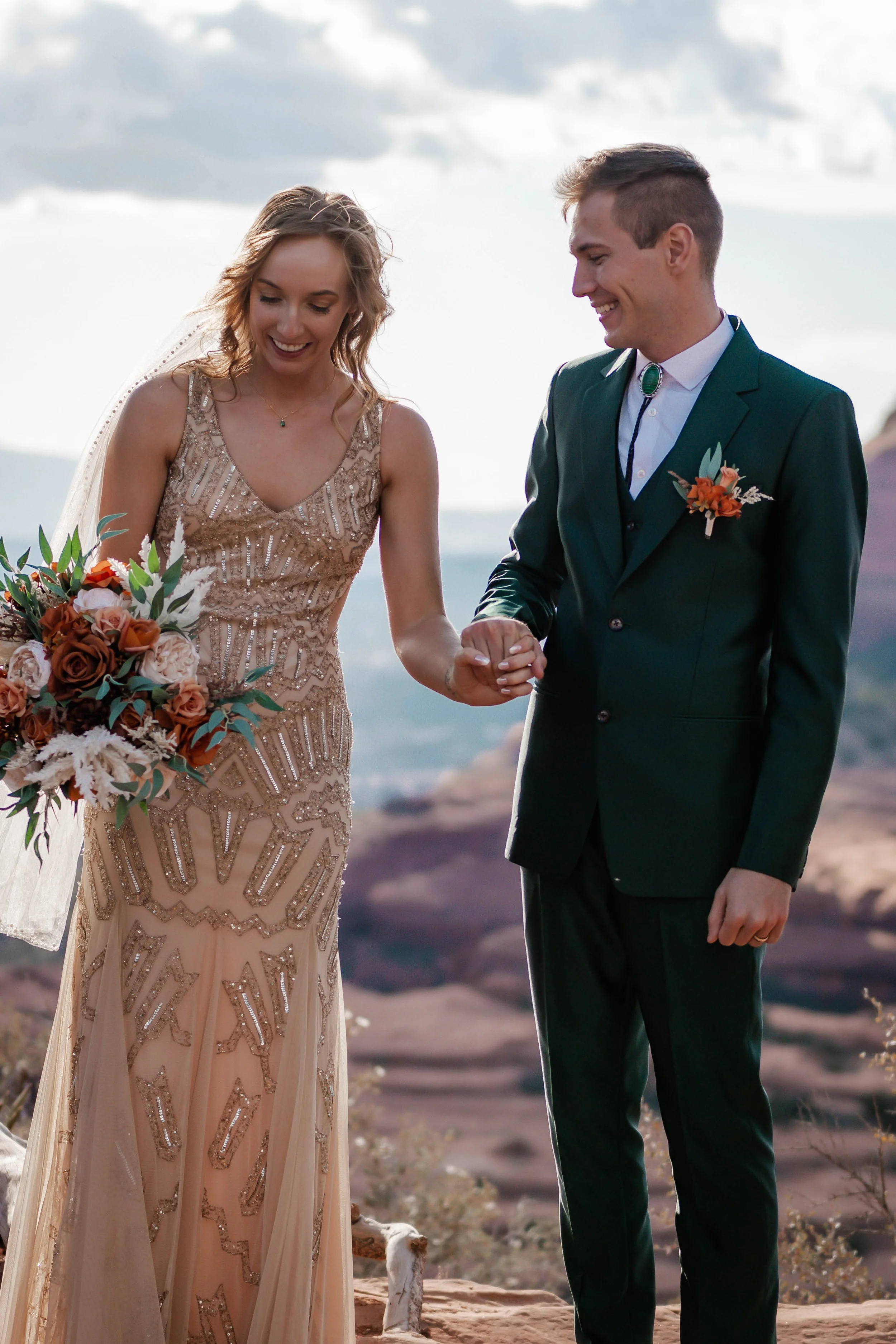 A bride and groom holding hands, standing outdoors on a rocky landscape with cloudy sky in the background, during their wedding ceremony.