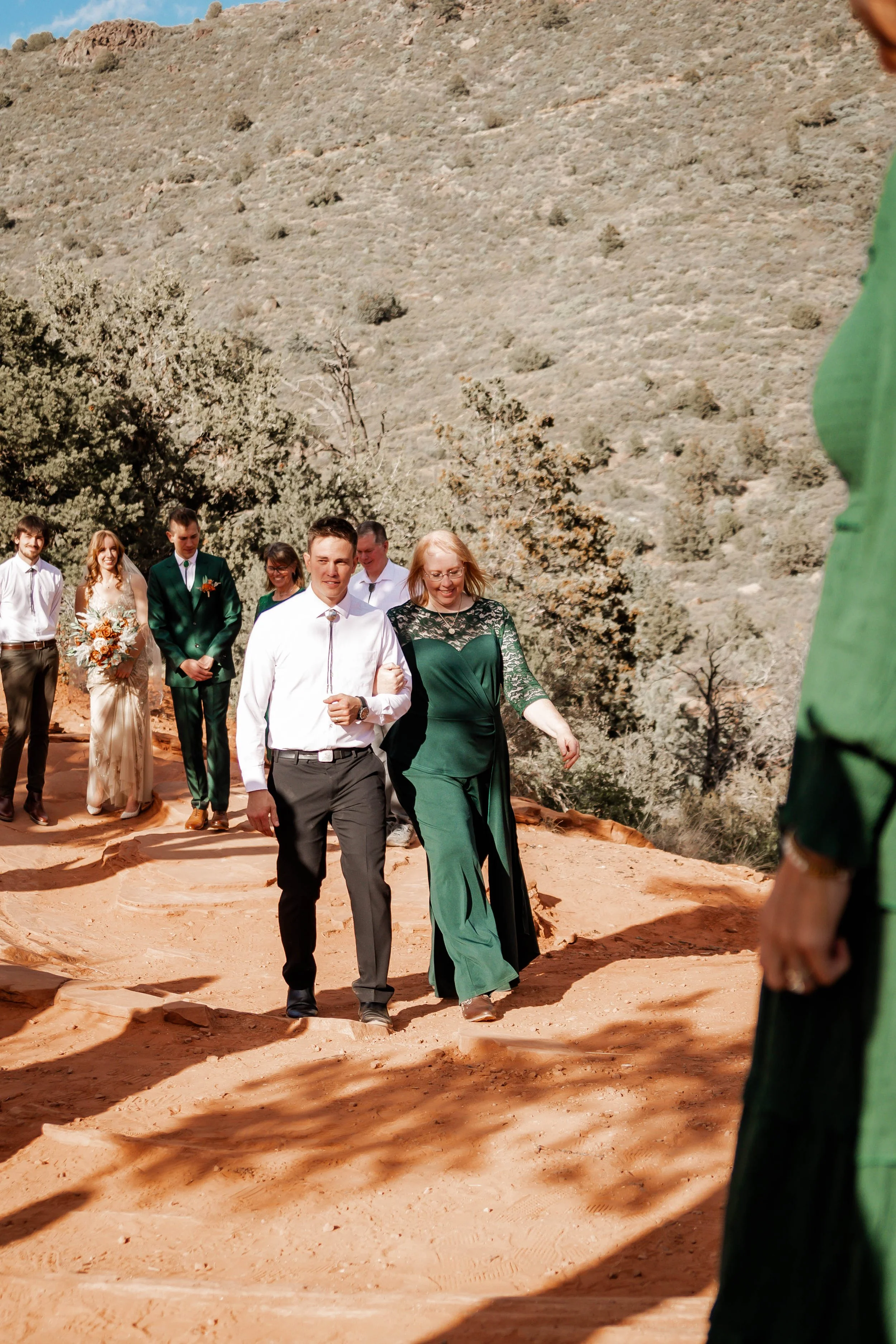 People walking outdoors on a dirt path, surrounded by nature and hills, likely during a wedding or special event.