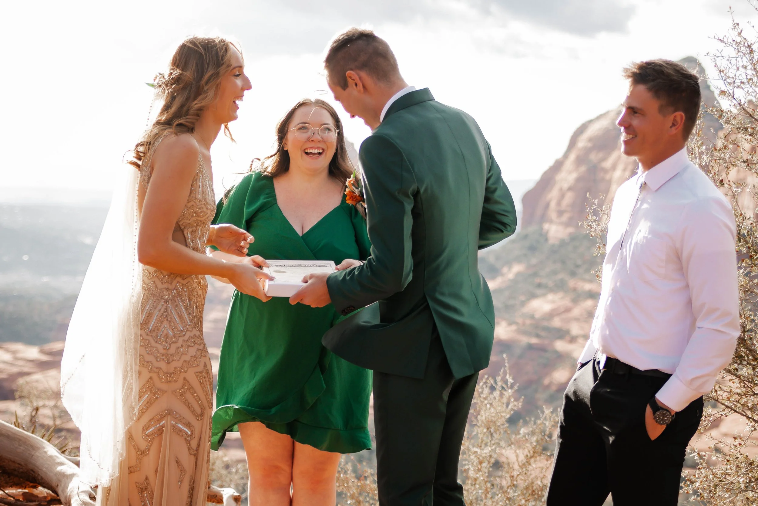 A wedding ceremony outdoors with four people, a bride in a gold dress, a groom in a green suit, a woman in a green dress, and a man in a white shirt. The couple exchanges vows in front of a scenic mountainous background.