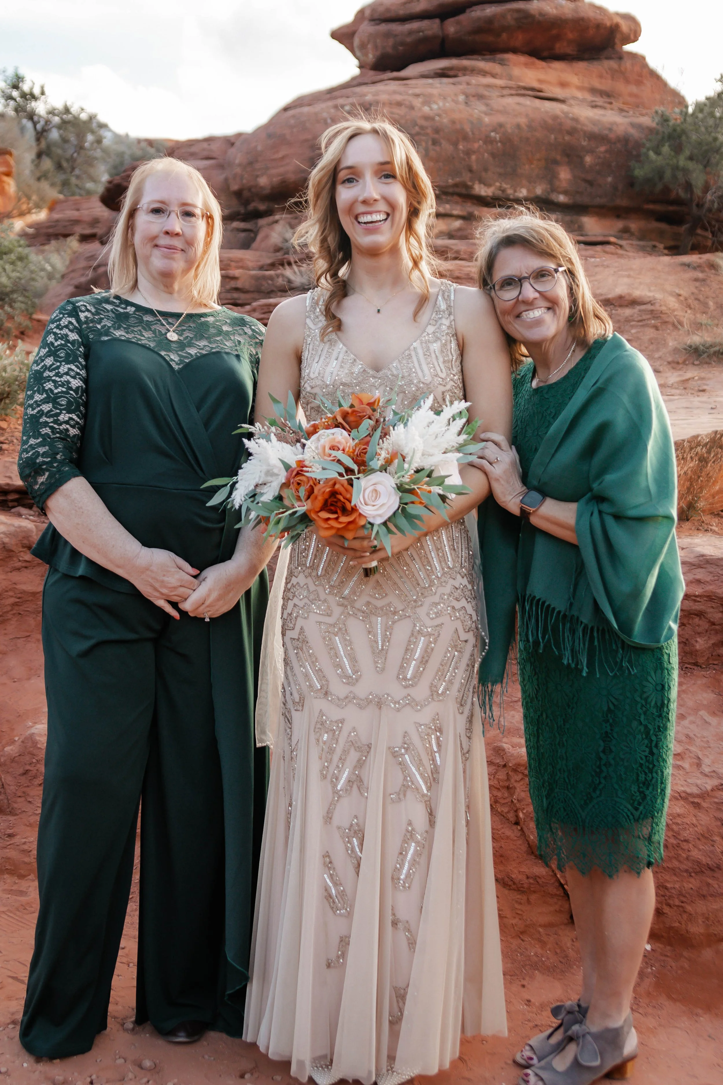Women standing outdoors in front of rocky terrain; one woman in a champagne-colored dress holding a bouquet, flanked by two women in green dresses.