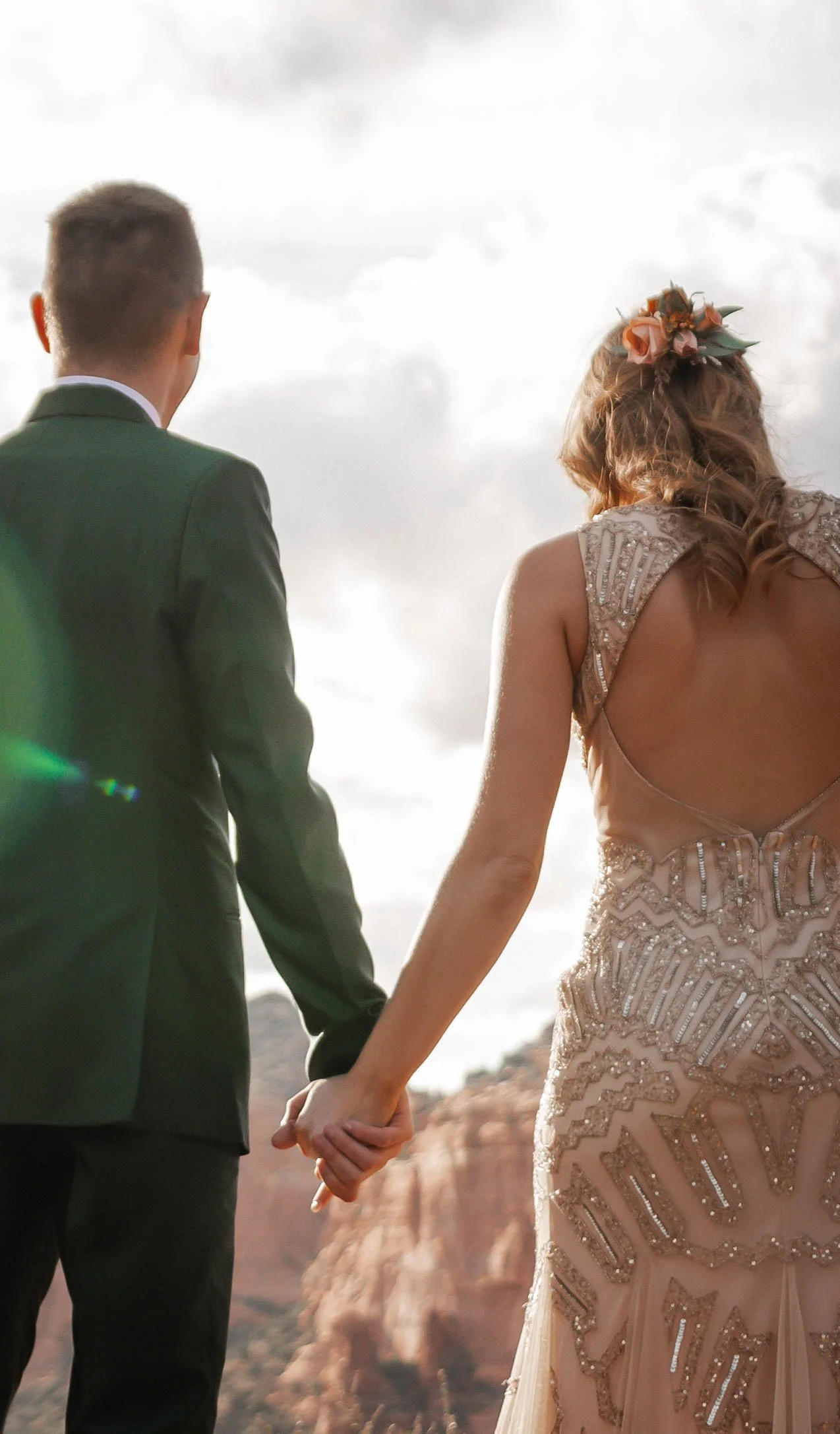 Couple holding hands and facing a rocky landscape, one wearing a suit and the other a beaded dress with floral headpiece, during a cloudy outdoor event.