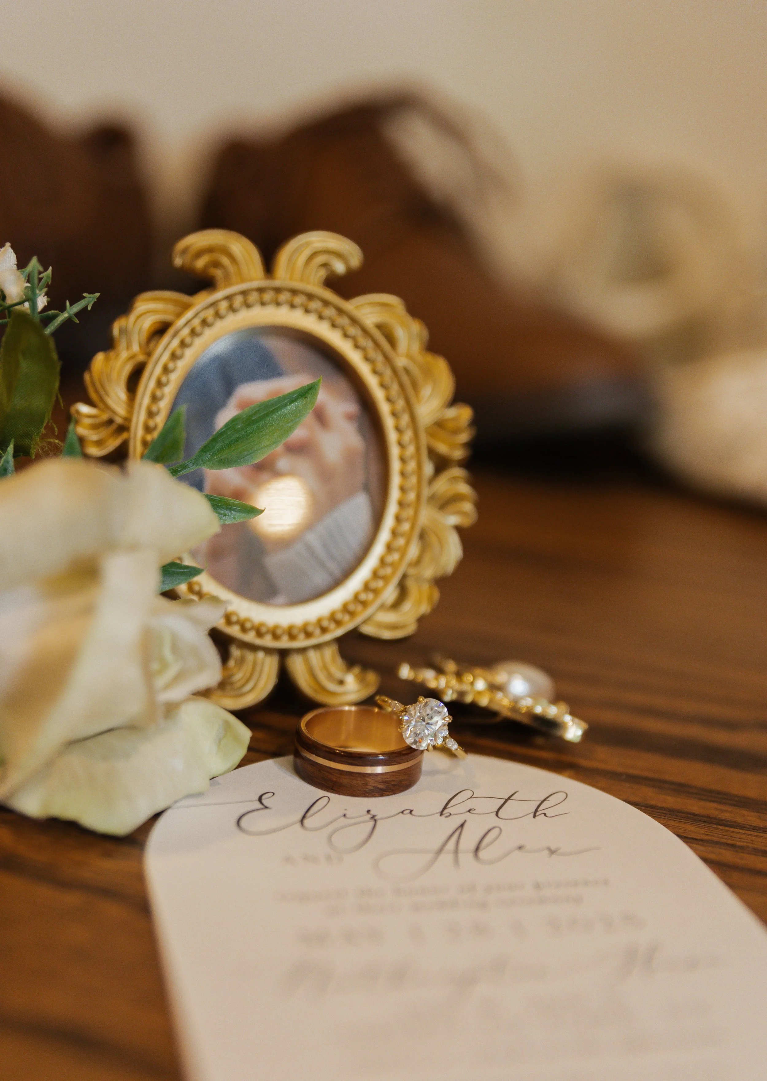 A wedding ring and an engagement ring placed on a piece of paper with calligraphy writing, positioned in front of a small ornate gold picture frame containing a blurred photograph of a couple. There are some white flowers and green leaves partially o