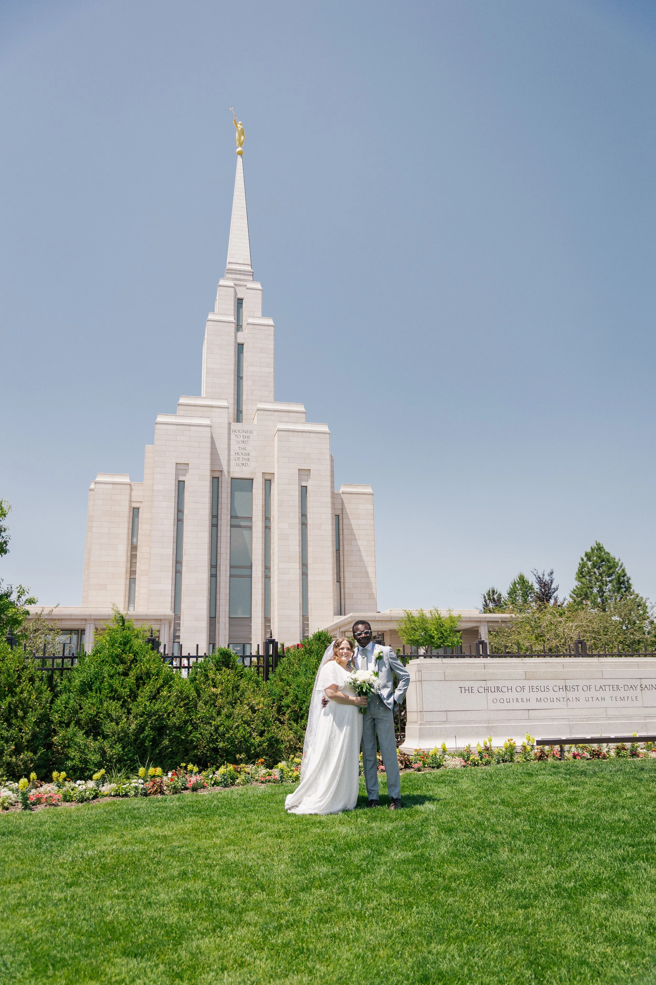Bride and groom standing together outside a church on their wedding day, with the church's steeple and a statue at the top visible in the background.
