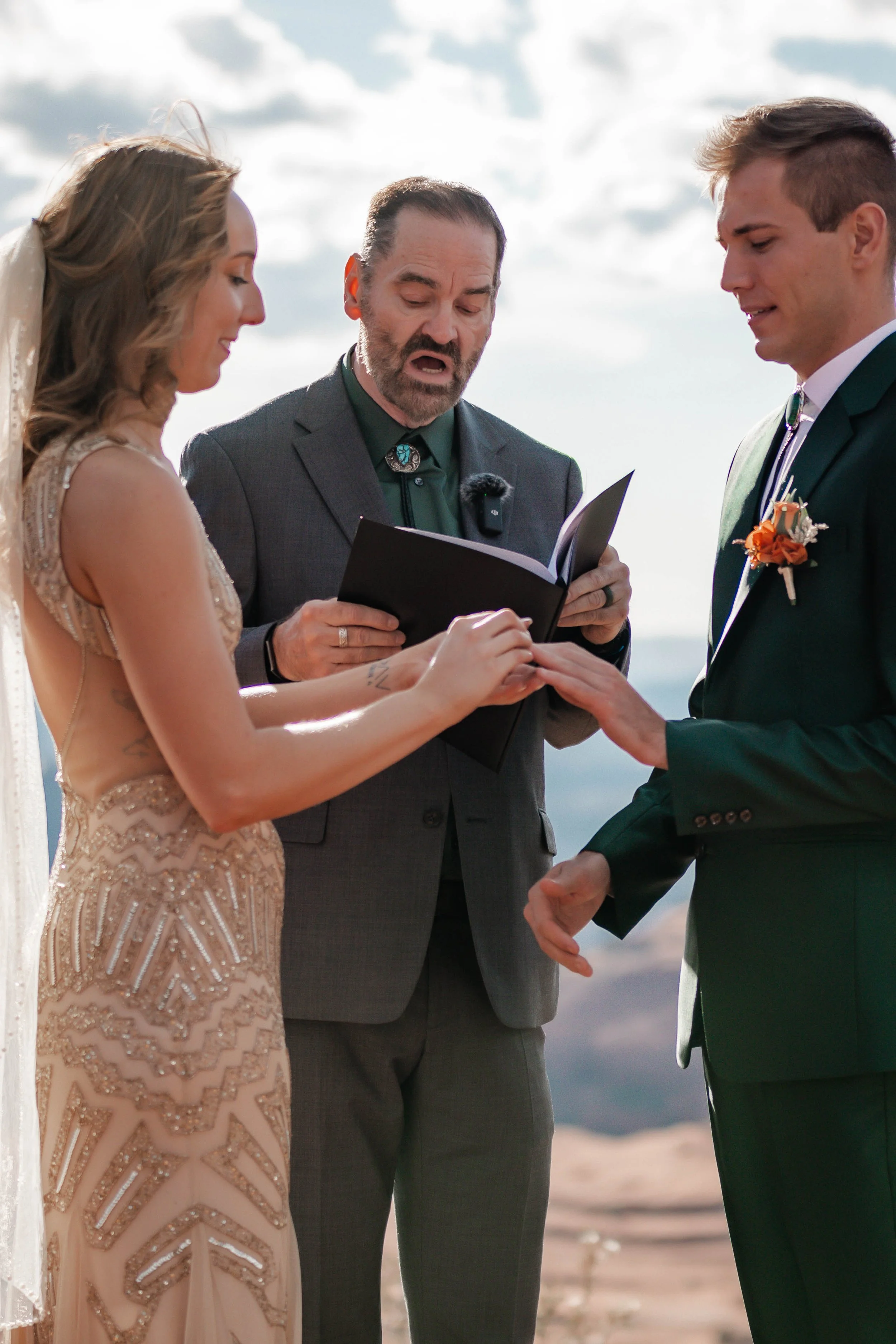 A wedding ceremony outdoors featuring a bride and groom exchanging rings, with an officiant reading from a book.