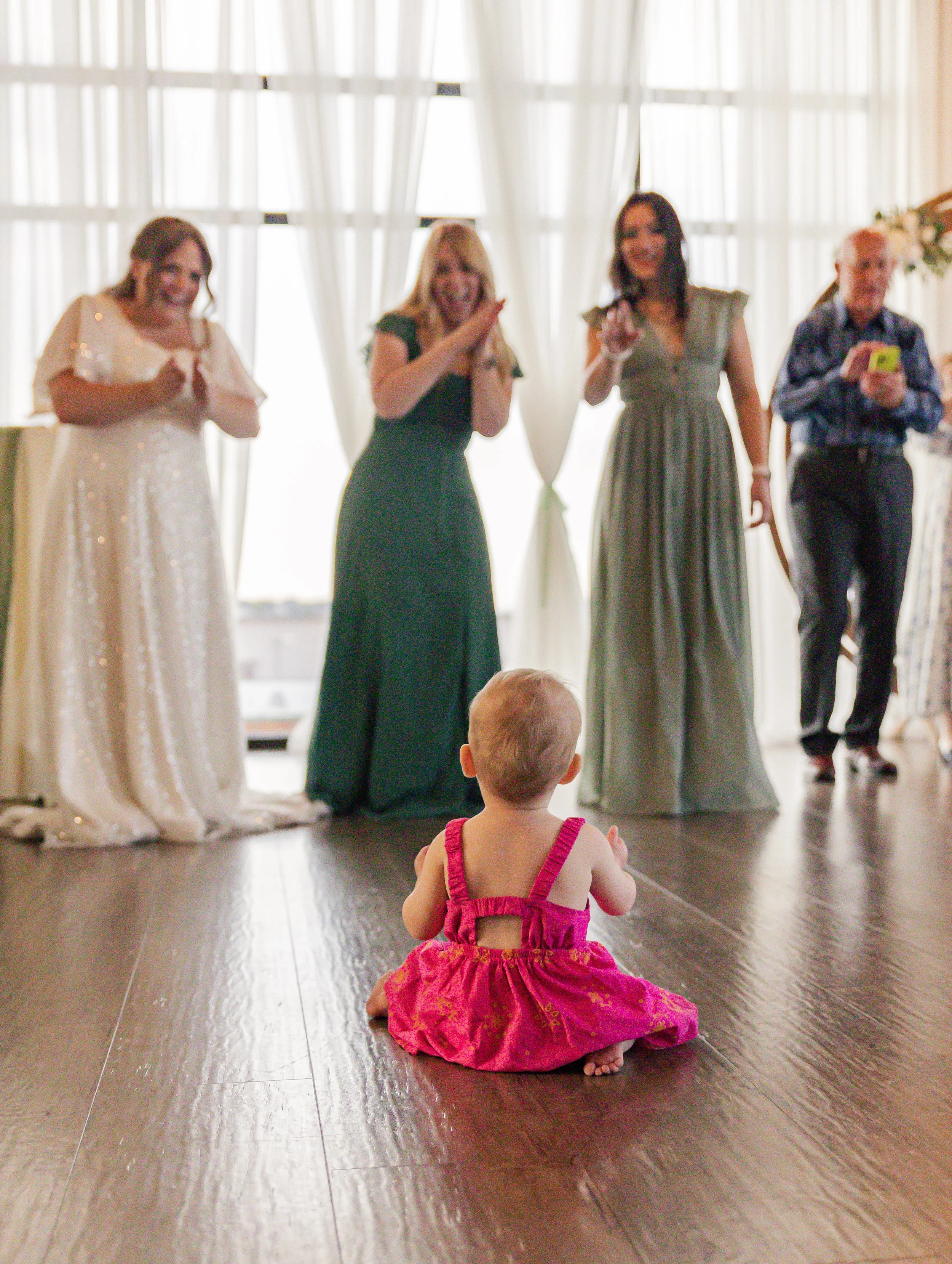 A baby girl in a pink dress sitting on a wooden floor facing a group of four adults who are standing in front of large windows with white curtains at a celebration or wedding event.