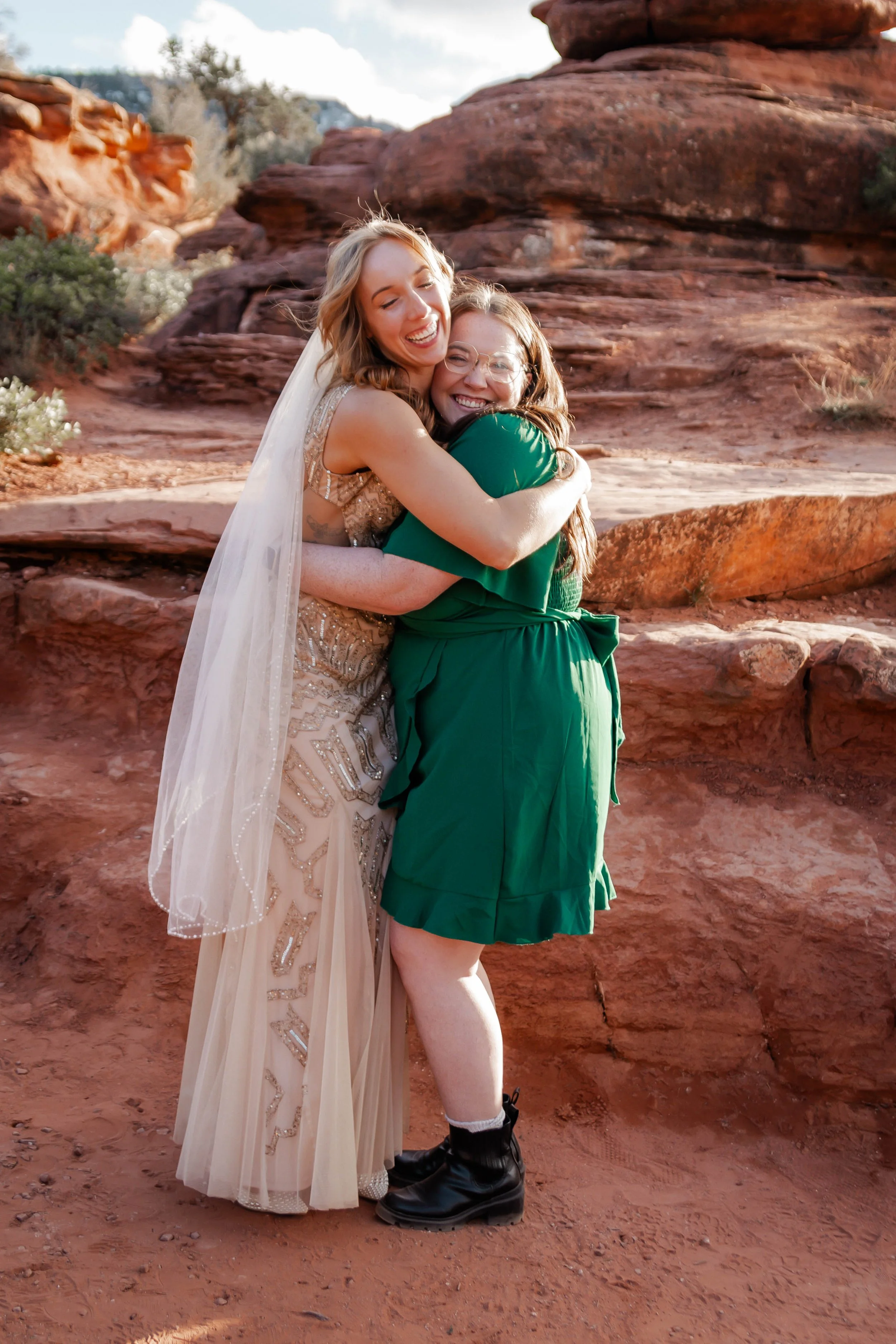 Two women hugging outdoors, one in a wedding dress with a veil, the other in a green dress, against a red rock landscape.