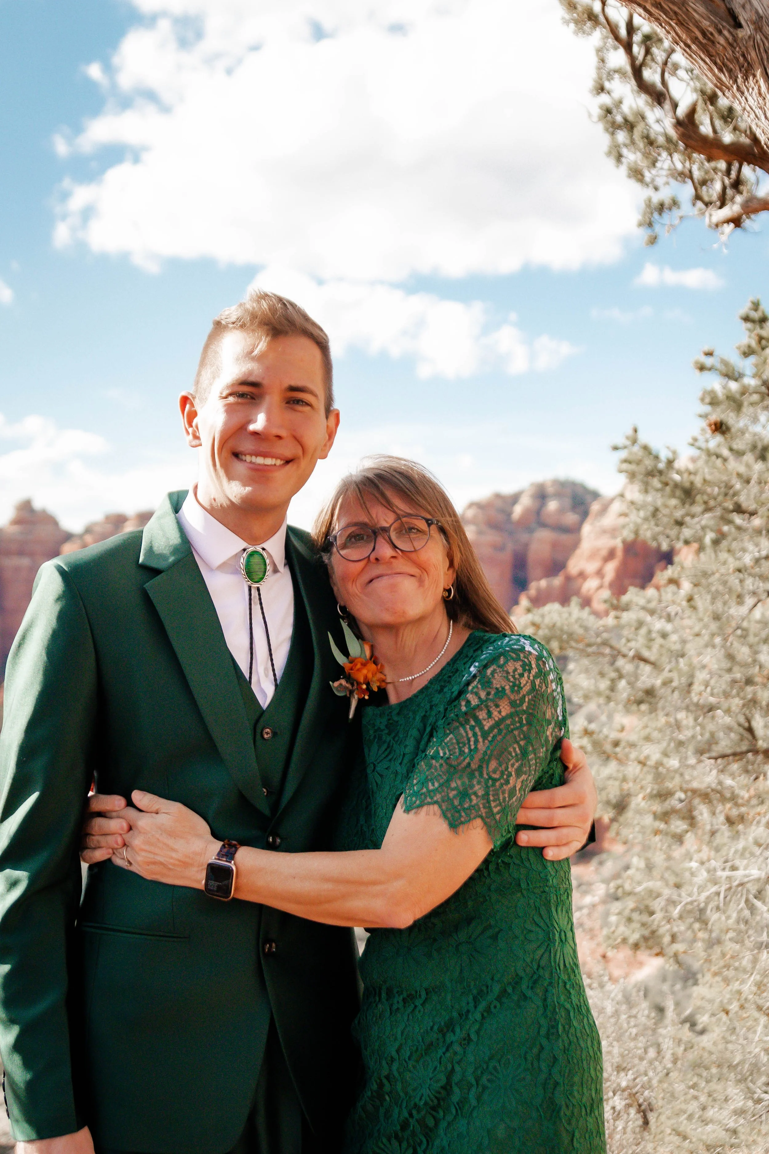 A young man in a green suit with a bolo tie and an older woman in a green lace dress hugging outdoors with red rock formations and trees in the background.