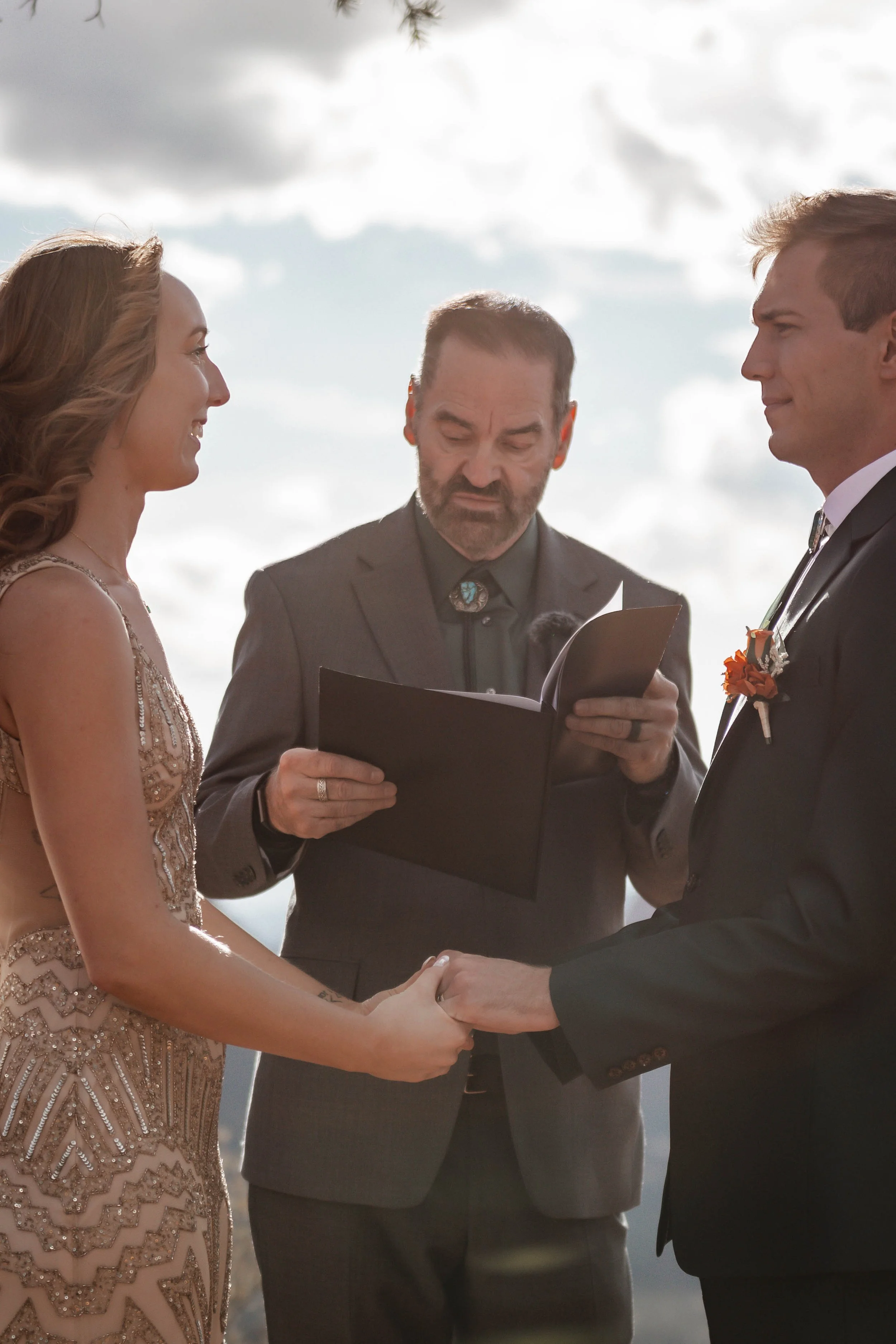 A couple getting married outdoors, holding hands and facing each other, with a officiant reading from a book.