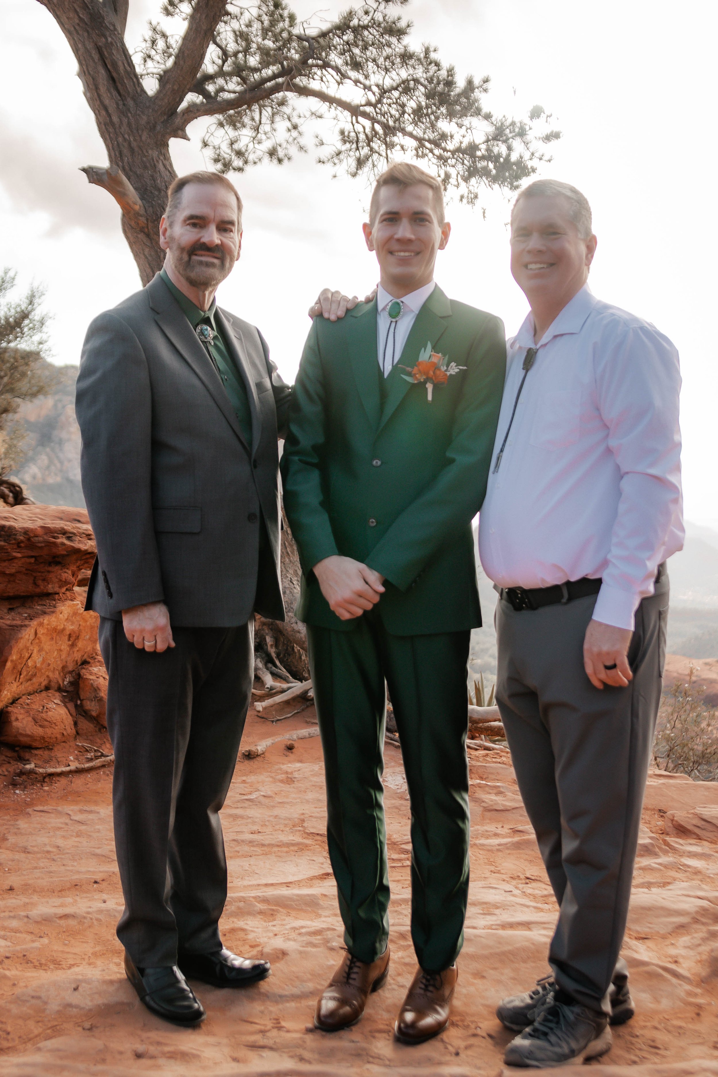 Three men standing outdoors on a rocky terrain with a tree behind them, at sunset. One man is wearing a dark gray suit, the second man, in the middle, is wearing a green suit with a boutonniere, and the third man is wearing a white shirt and gray tro