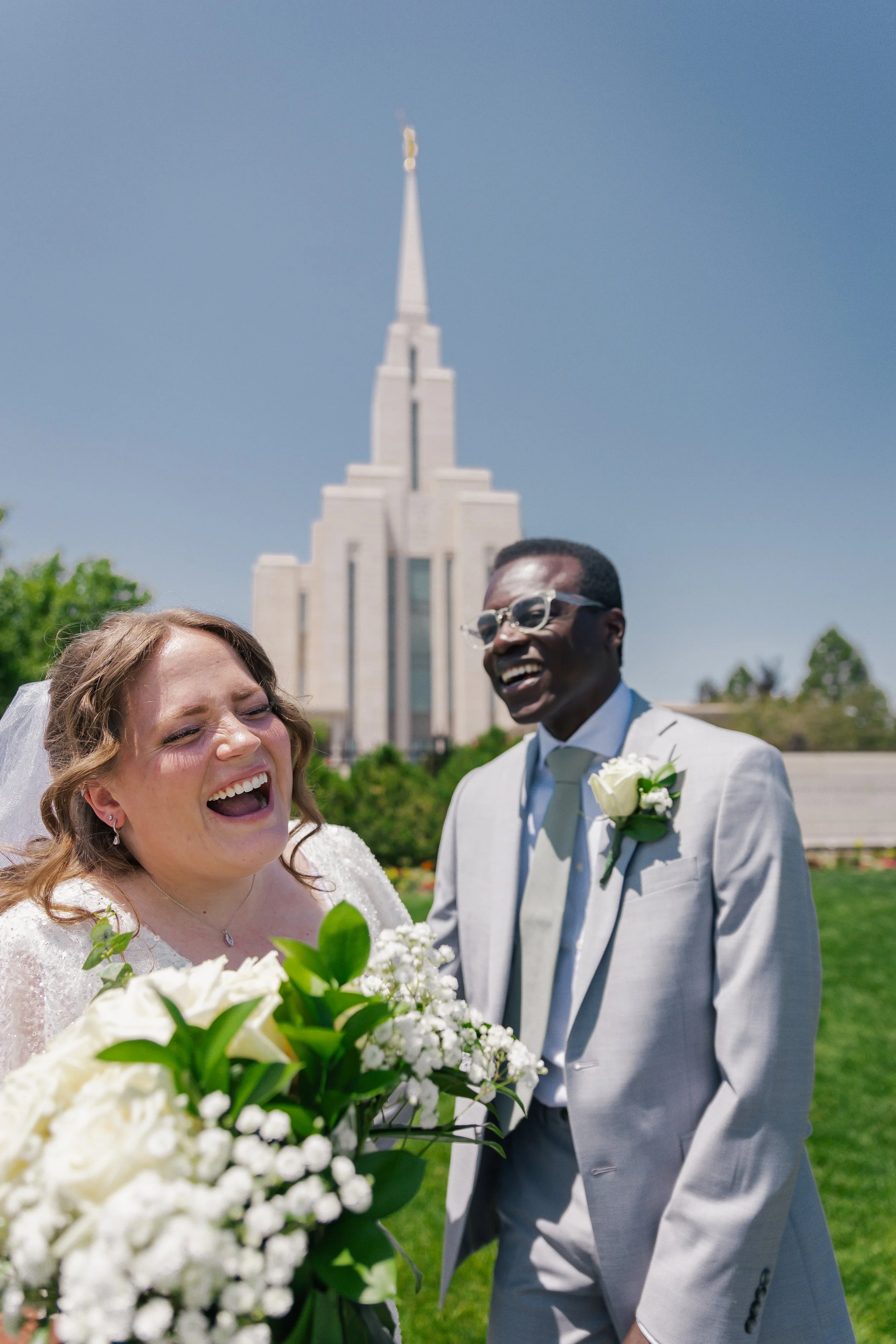 Two people in wedding attire laughing outdoors with oquirrh mountain temple in the background.