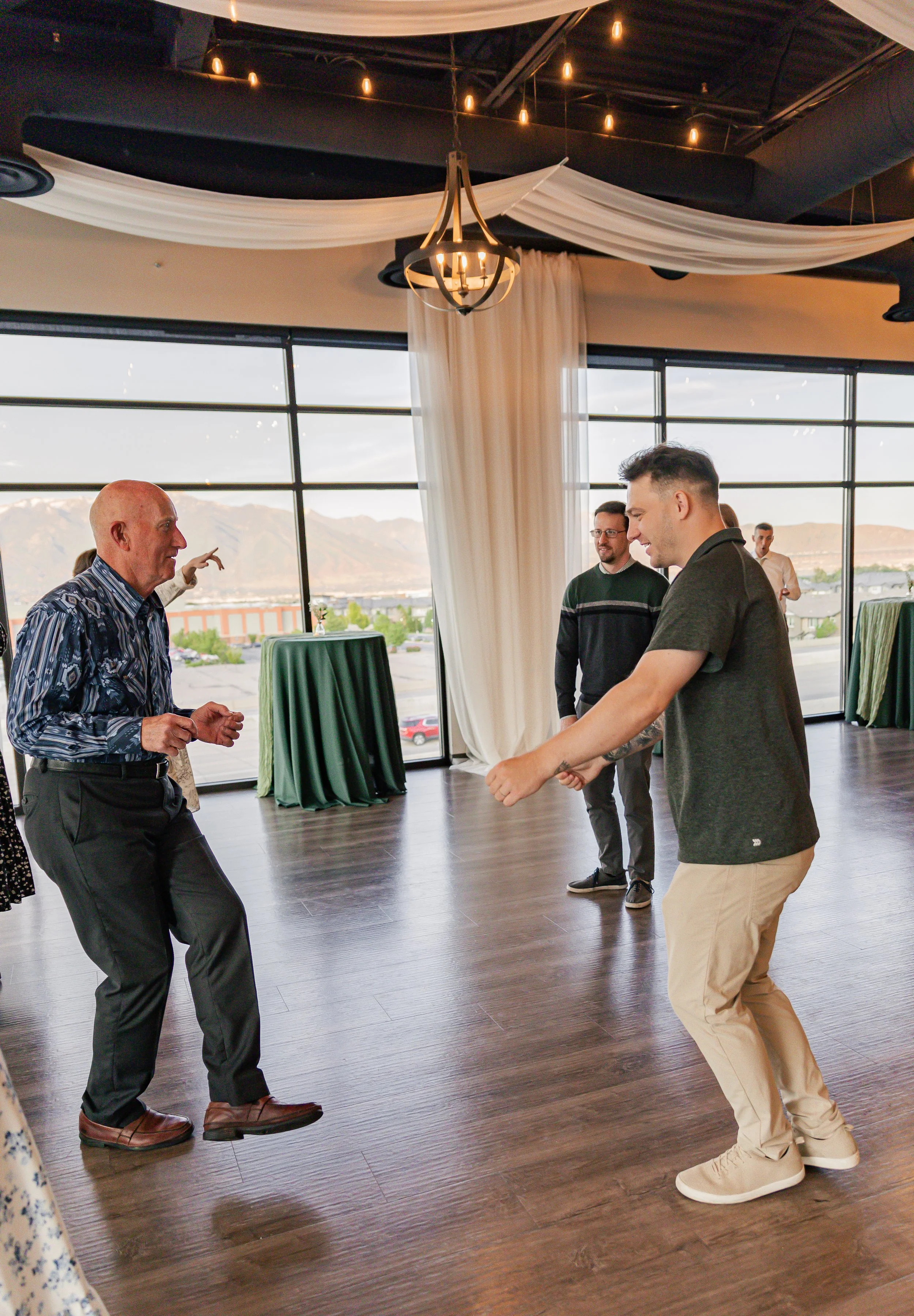 People dancing and enjoying themselves at a celebration in a venue with large windows, white drapery, and a view of mountains.
