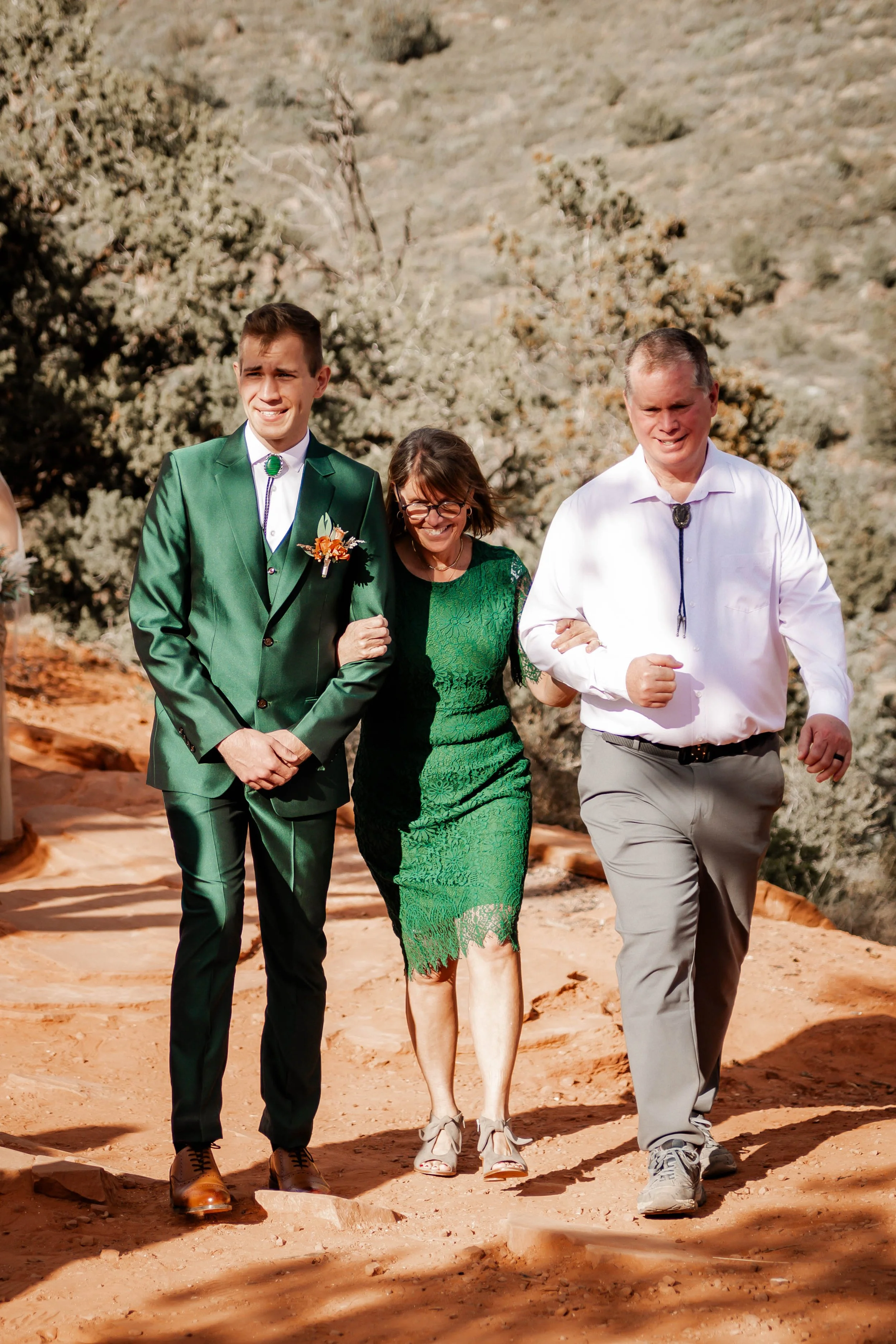 Three people walking outdoors on a dirt path, two men and one woman, with desert vegetation and hills in the background.