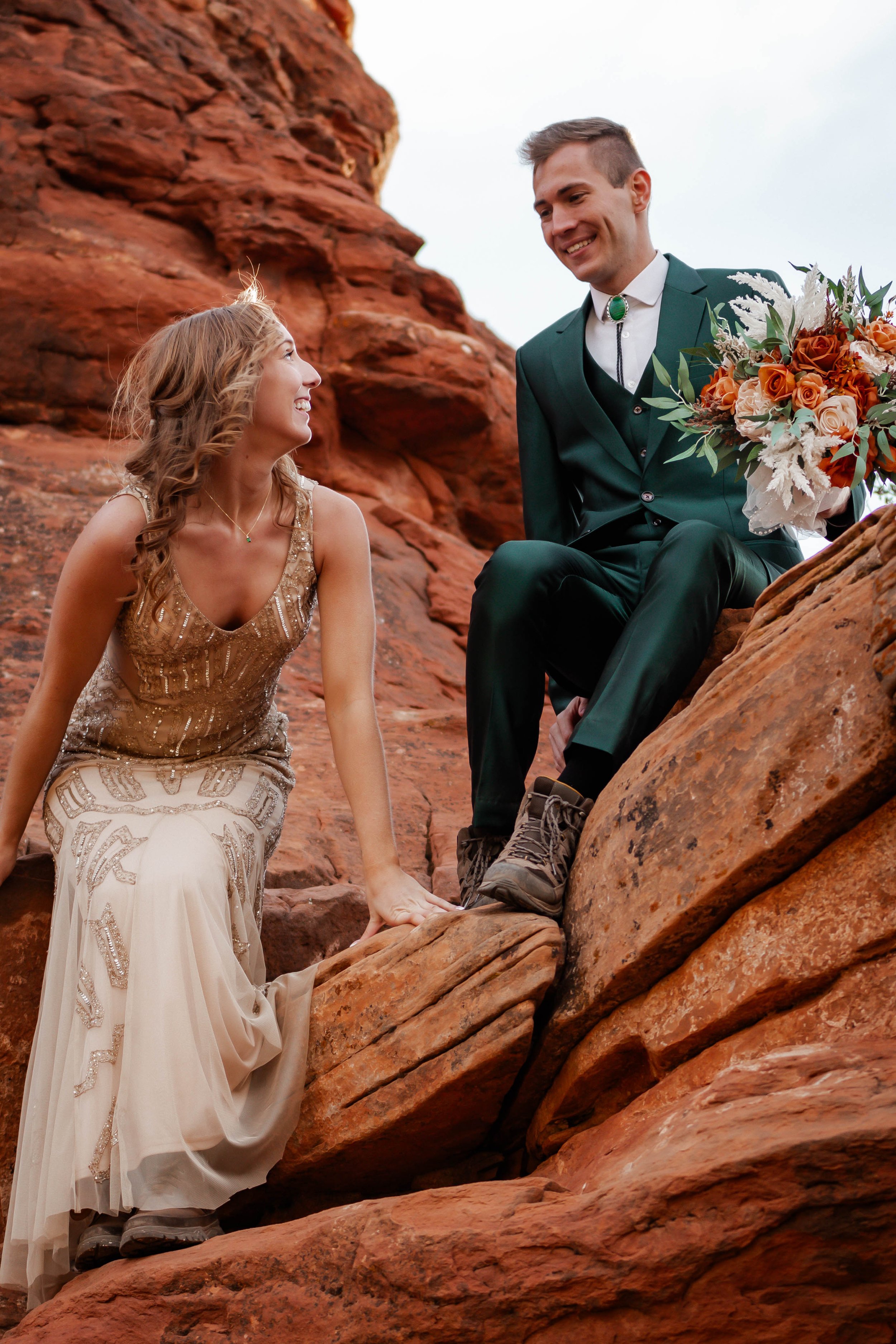 A couple dressed in wedding attire on a rocky outdoor landscape. The woman wears a beige dress with intricate beading and lace, while the man is in a green suit with a white shirt and a bolo tie, holding a bouquet of flowers.