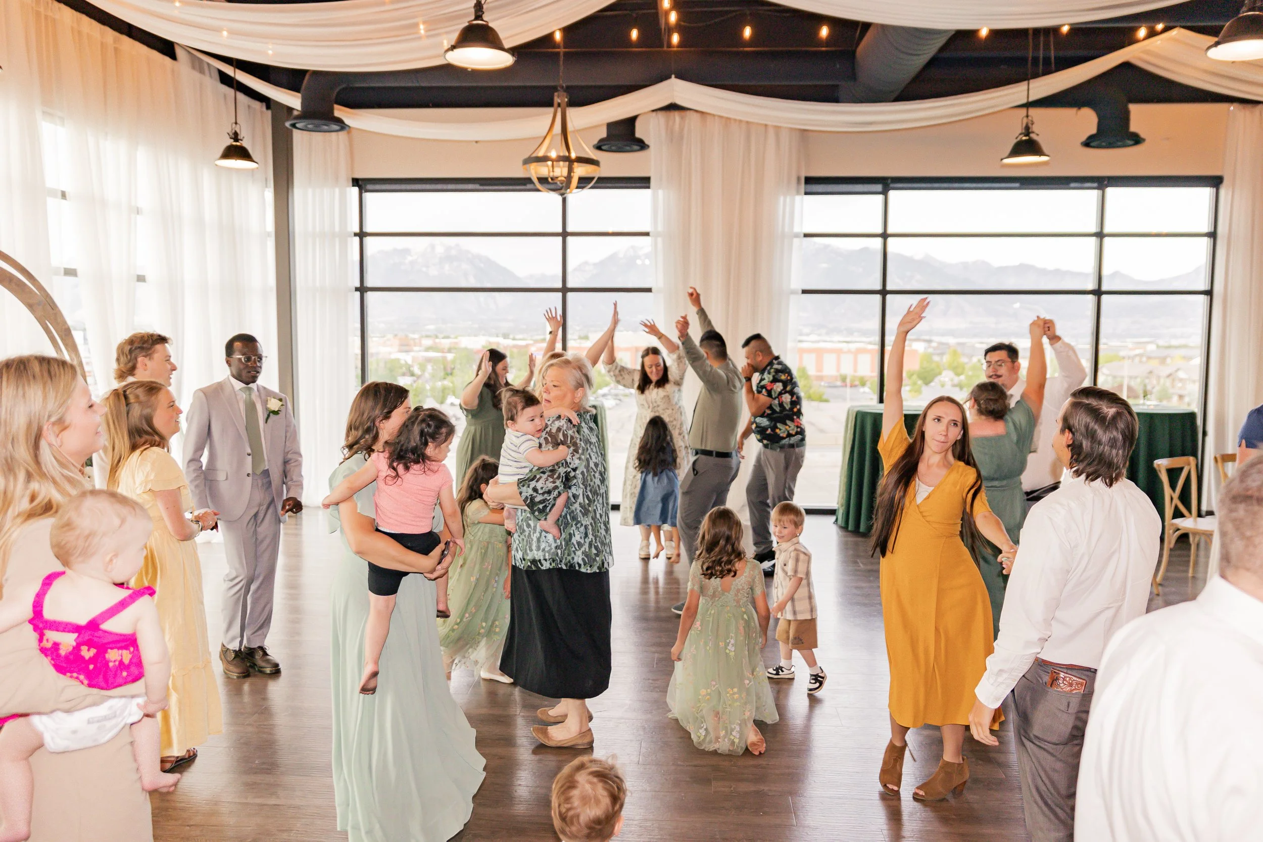 group of people dancing and celebrating at an indoor event, with children and adults, in a decorated hall with large windows and mountain views.