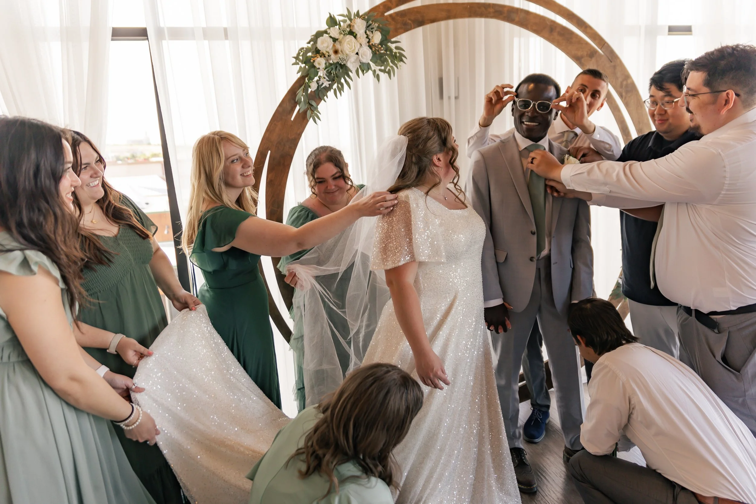 Bride and groom surrounded by friends at a wedding, with friends adjusting glasses and helping with the dress in a decorated indoor space.