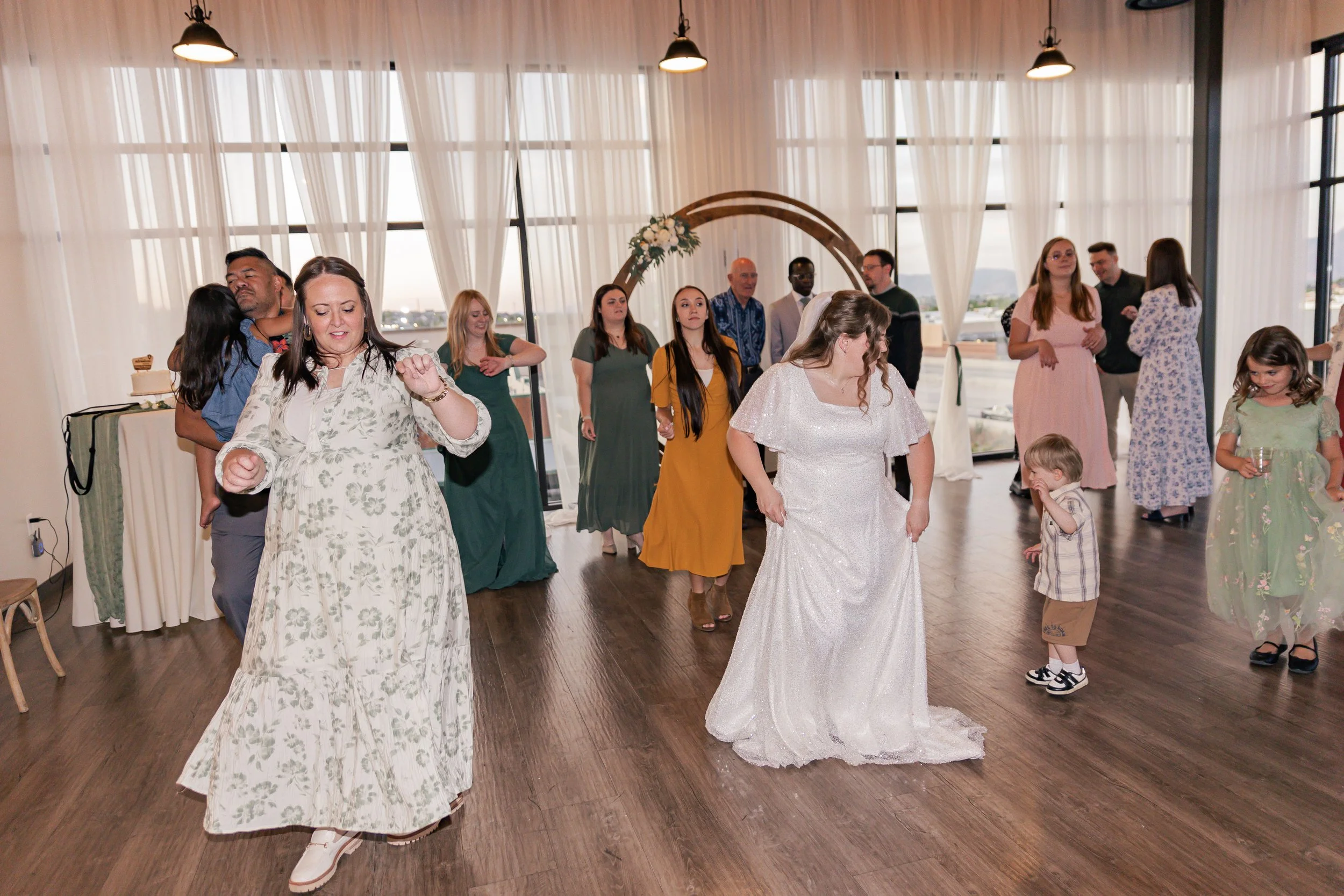 People dancing and socializing at a wedding reception in a bright, modern venue with large windows and sheer curtains.