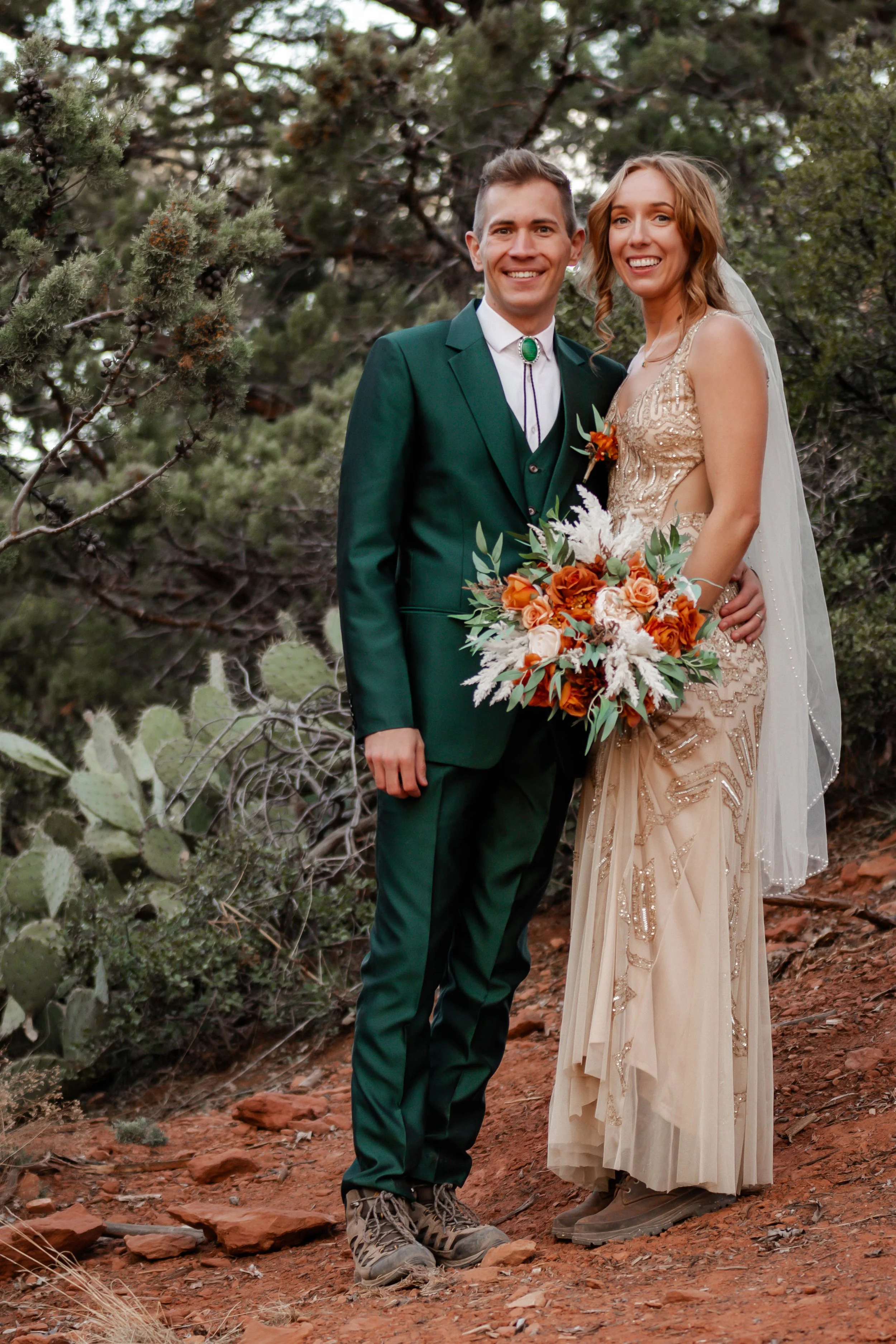 A smiling couple in wedding attire standing outdoors in a forested area with desert plants, holding a bouquet of orange and white flowers.
