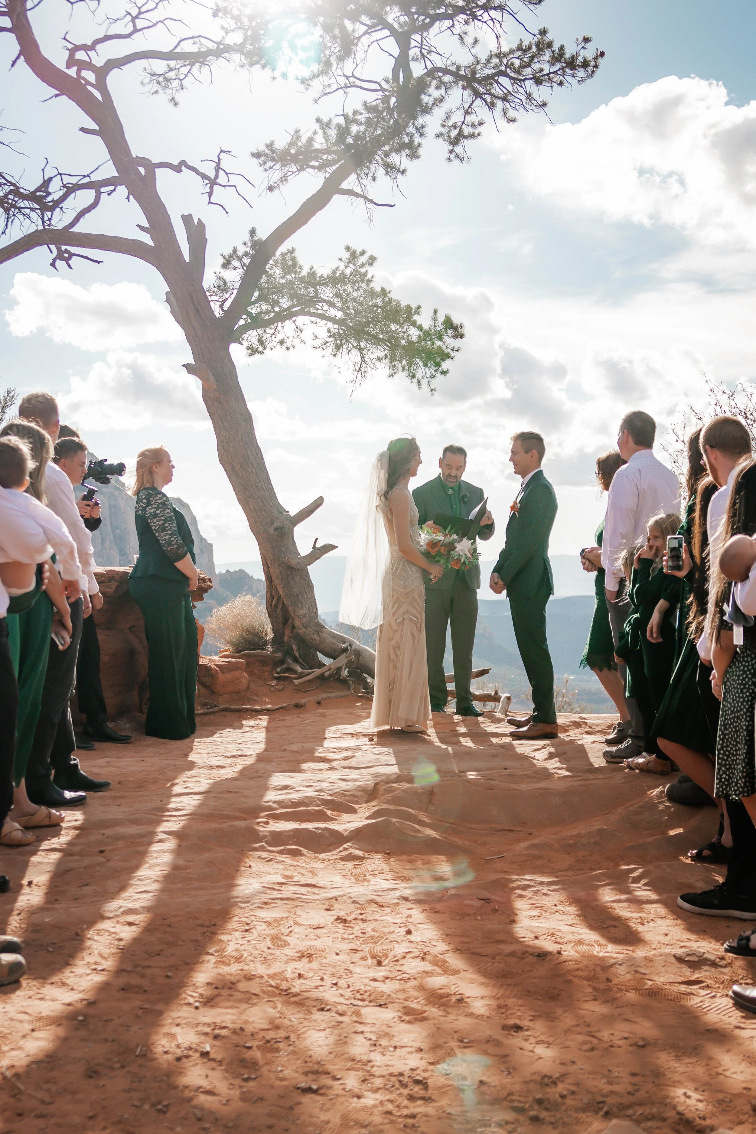 A wedding ceremony taking place outdoors with a bride and groom standing under a large tree, surrounded by guests on a sunny day with scattered clouds, in a scenic mountainous area.