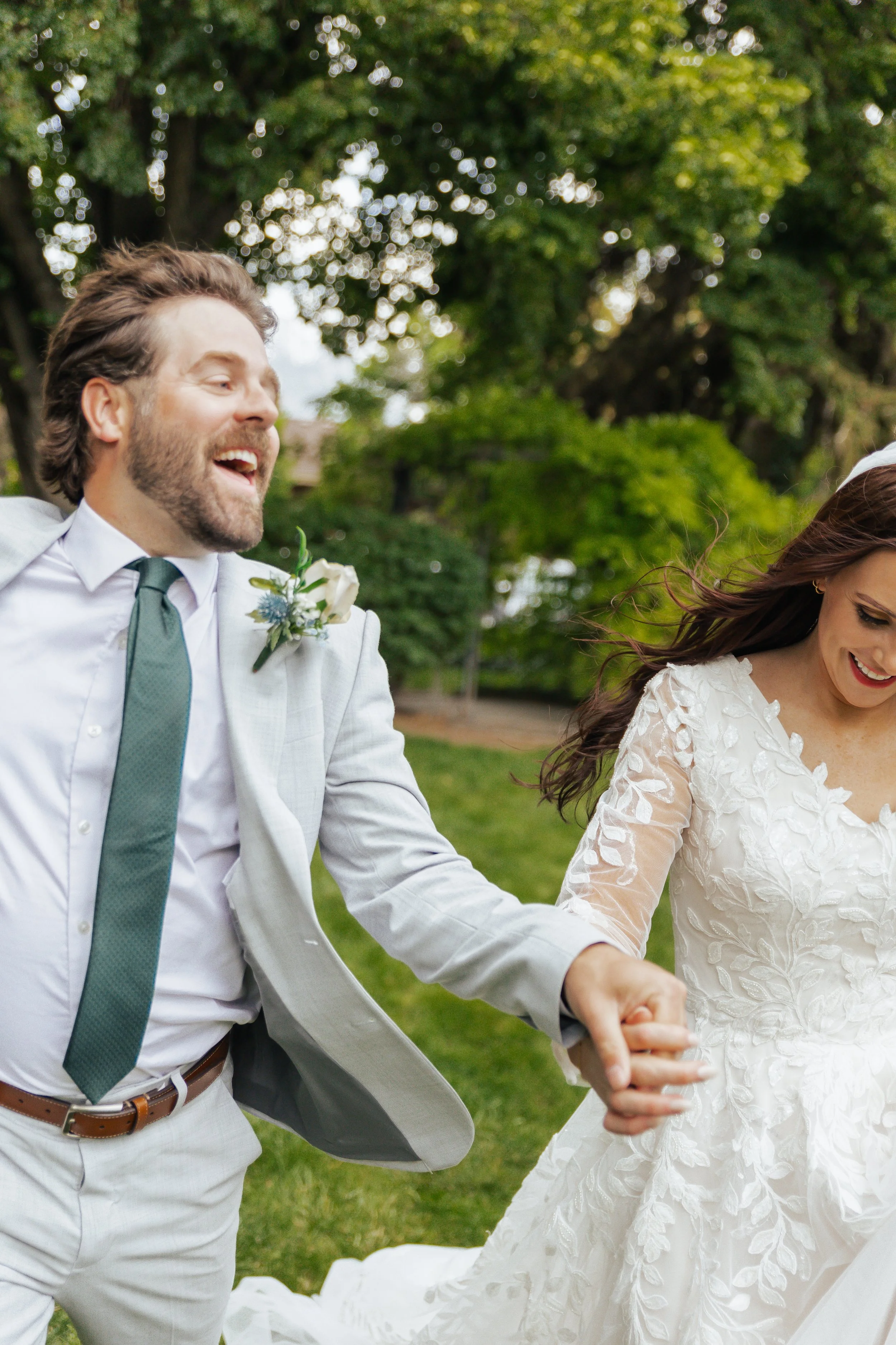A happy bride and groom holding hands and dancing outdoors on their wedding day, with trees and greenery in the background.