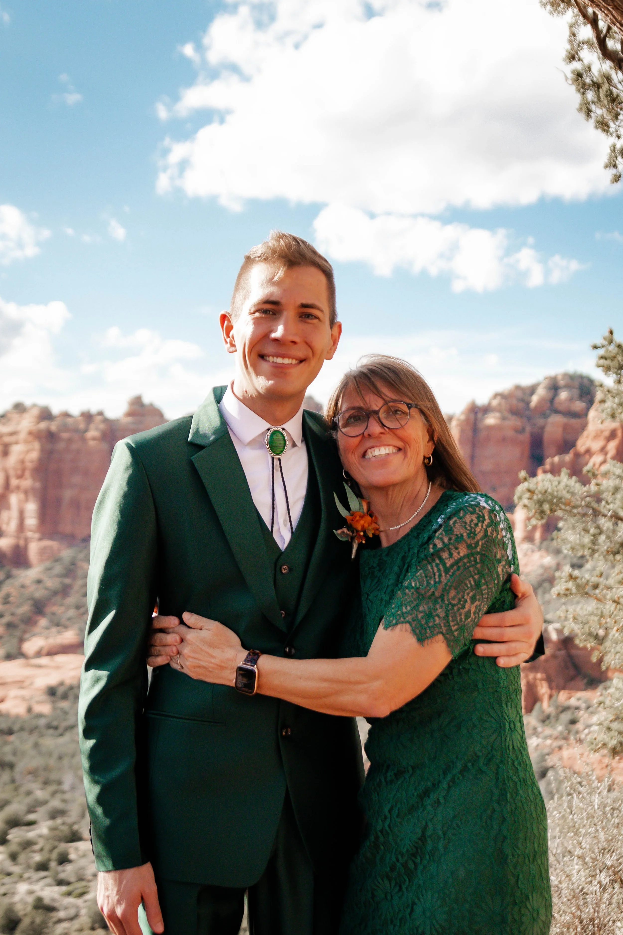 A young man and an older woman are smiling and hugging outdoors with red rock formations and a blue sky with clouds in the background.