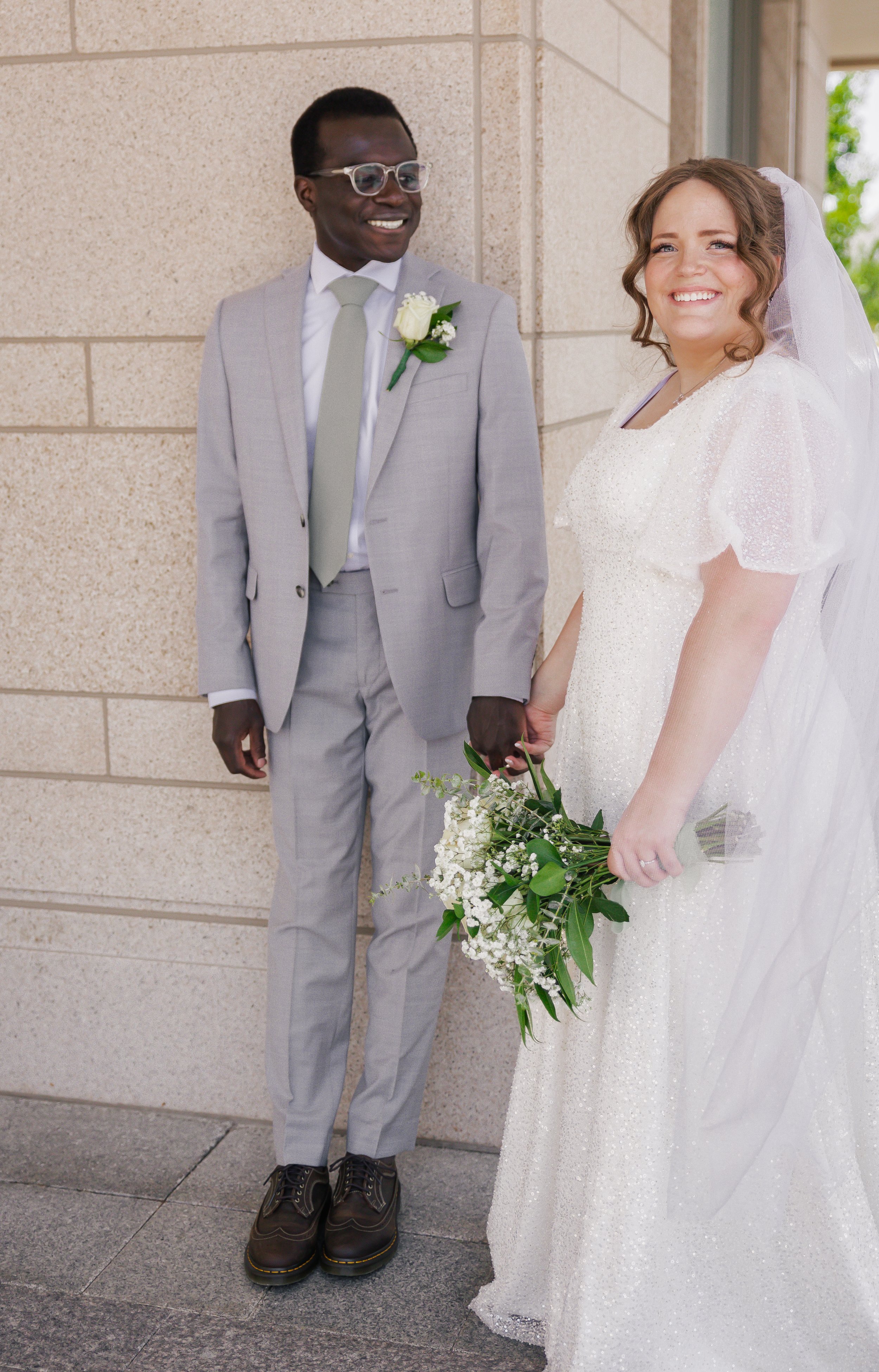 A smiling bride and groom holding hands outside a building. The groom is wearing a light gray suit with a white shirt, gray tie, glasses, and a white boutonniere. The bride is in a white wedding dress with puff sleeves, holding a bouquet of white flo