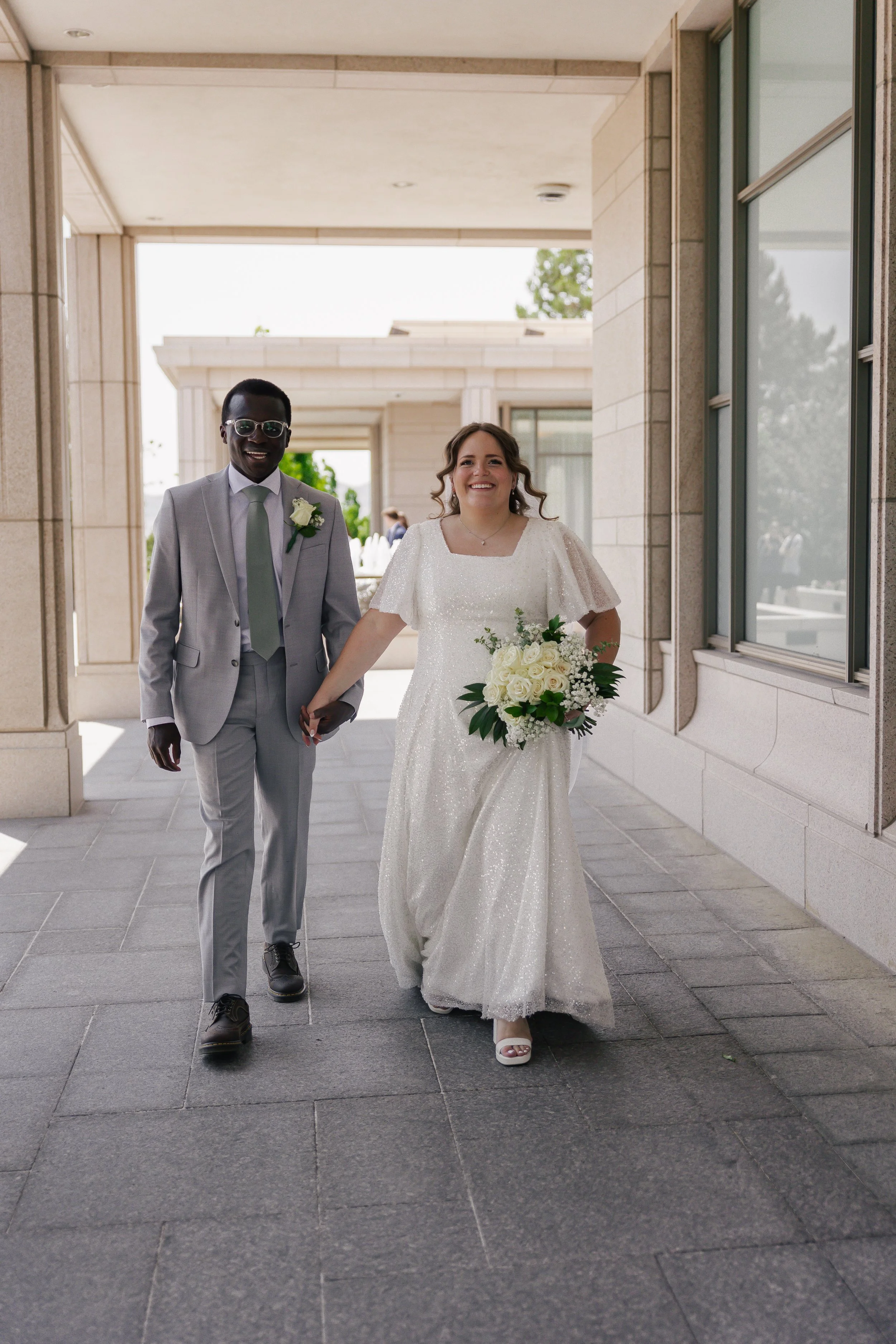 A smiling bride and groom walking hand in hand on their wedding day, outside a building with large windows and stone walls, the bride holding a bouquet of white roses and greenery.