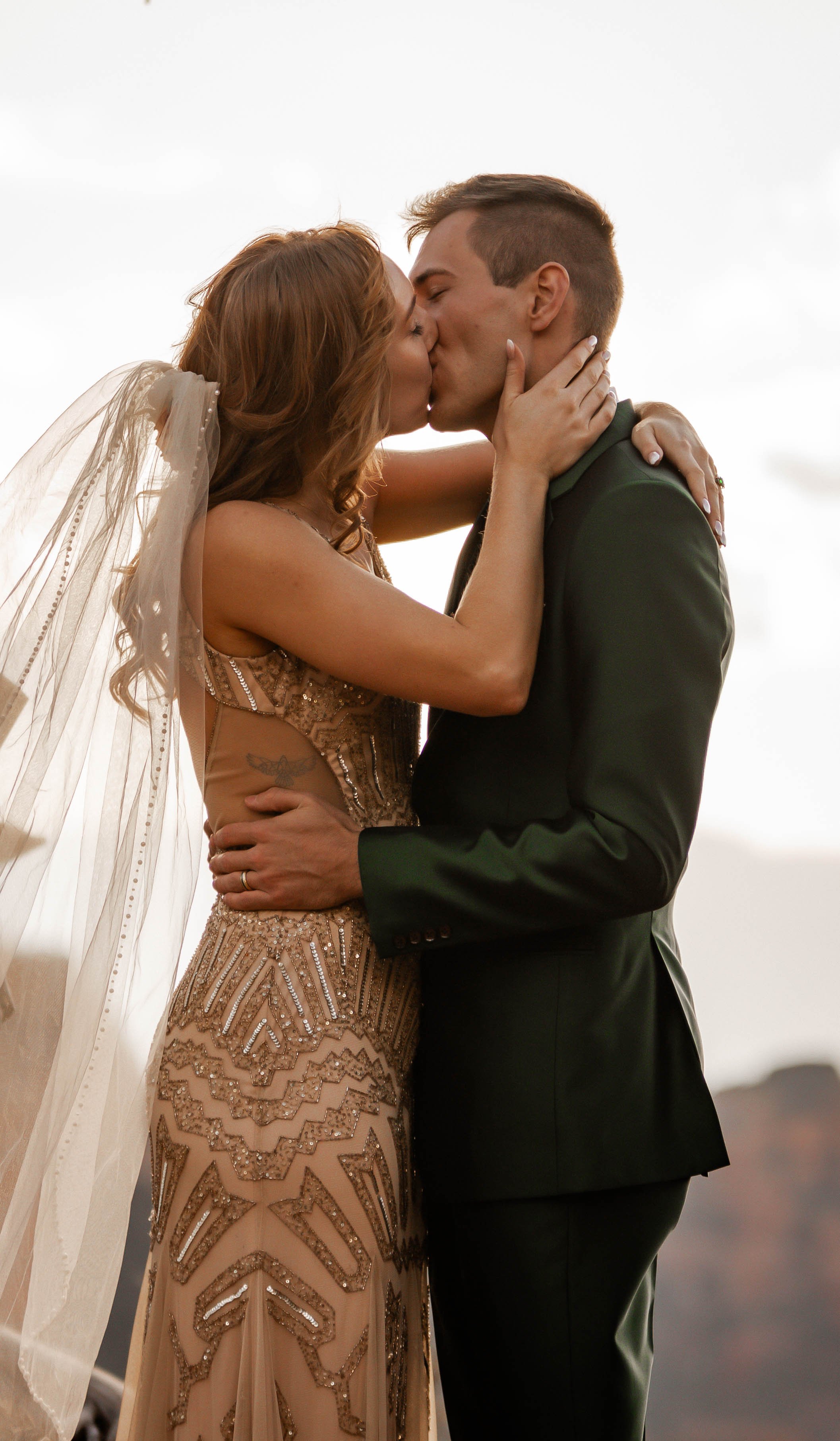 A couple sharing a kiss at sunset, the woman wearing an intricate, beaded gown with a veil, and the man in a dark suit.