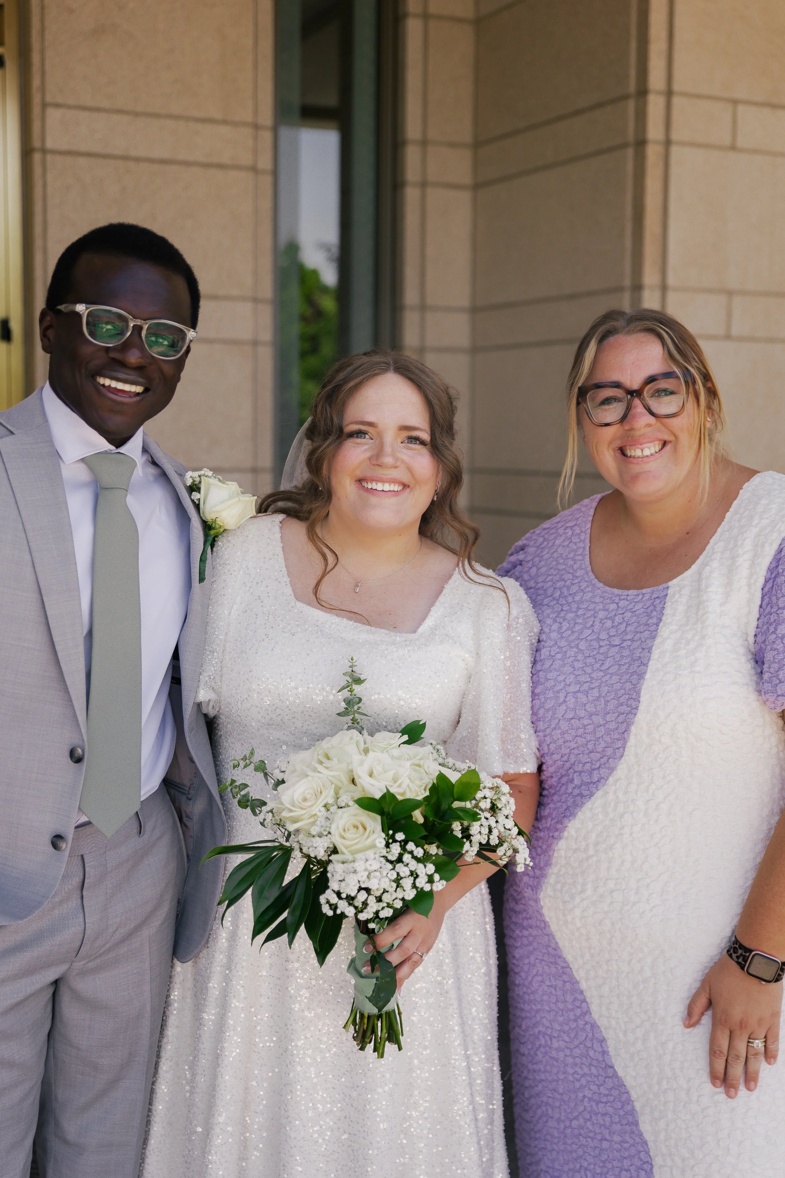 A bride in a white wedding dress holding a bouquet stands between a groom in a light gray suit and woman in a purple and white dress, outdoors near a building with beige stone walls.