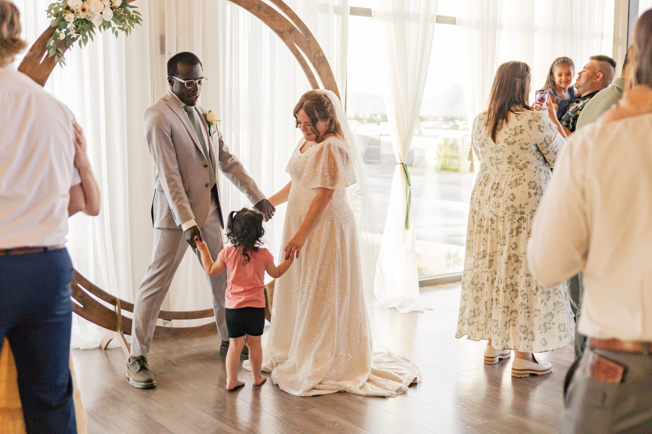A wedding ceremony scene with a bride and groom holding hands, surrounded by guests. The bride is wearing a white dress, and the groom is in a light gray suit. A small child stands between them, holding the bride's hand. Guests are taking photos and 