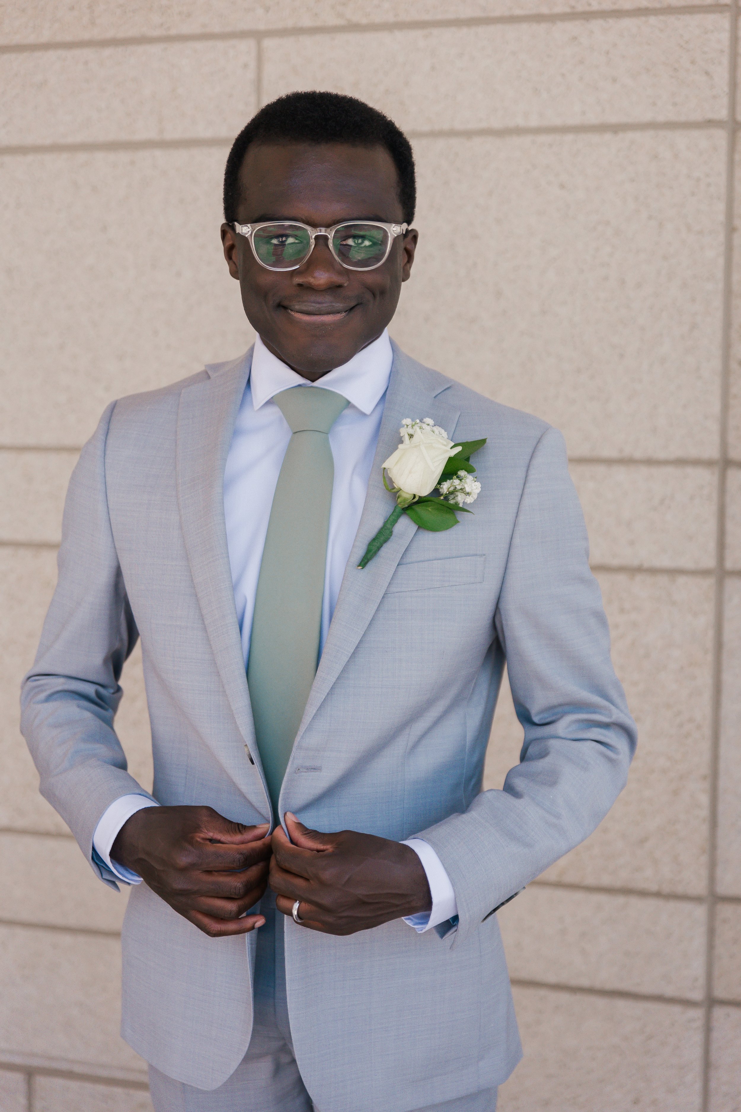 A young man in a light gray suit with a white shirt and pale green tie, smiling, standing in front of a beige brick wall. He has glasses and a white boutonniere on his lapel.