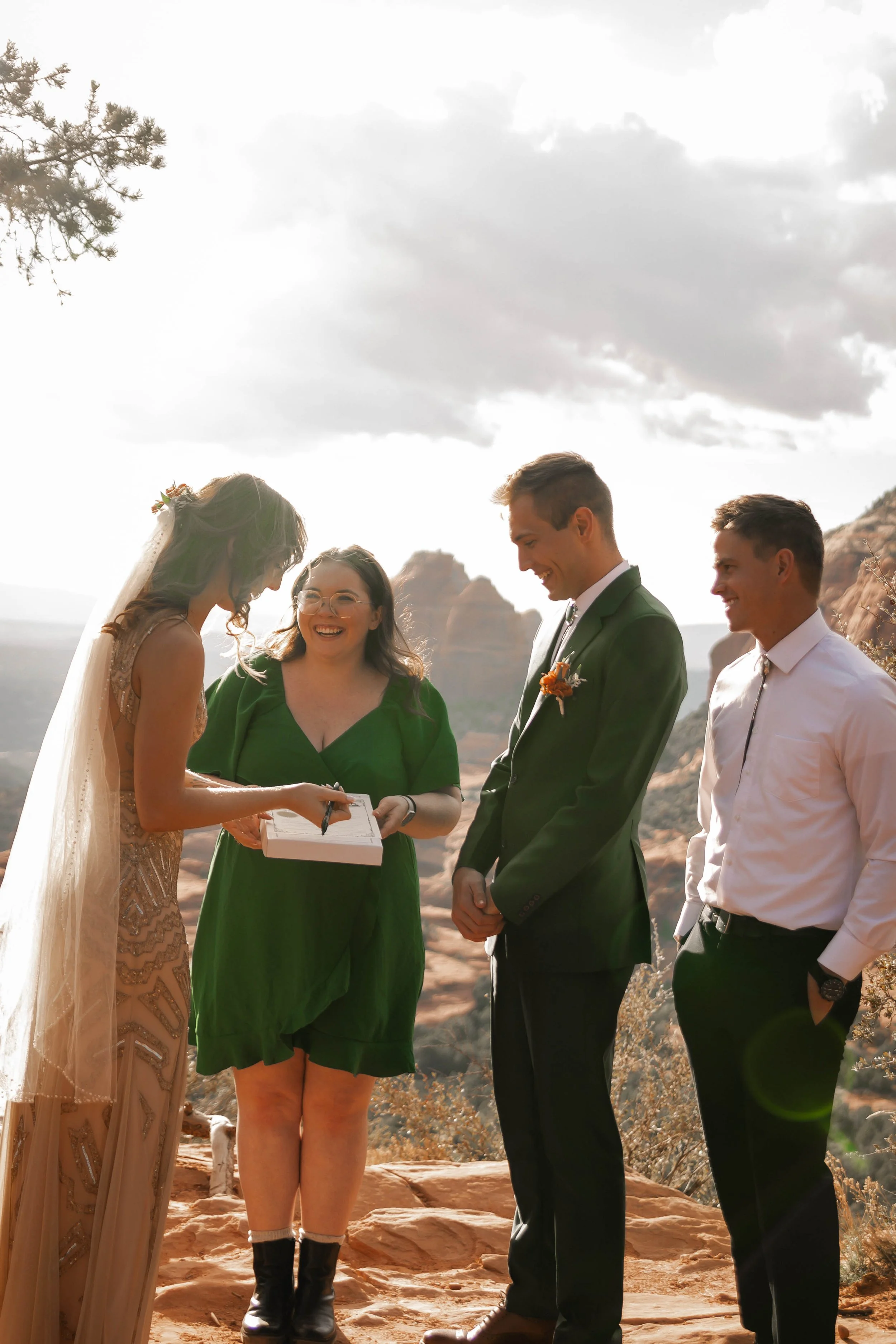 A wedding ceremony outdoors with four people, two men and two women, exchanging vows with a scenic desert landscape in the background.
