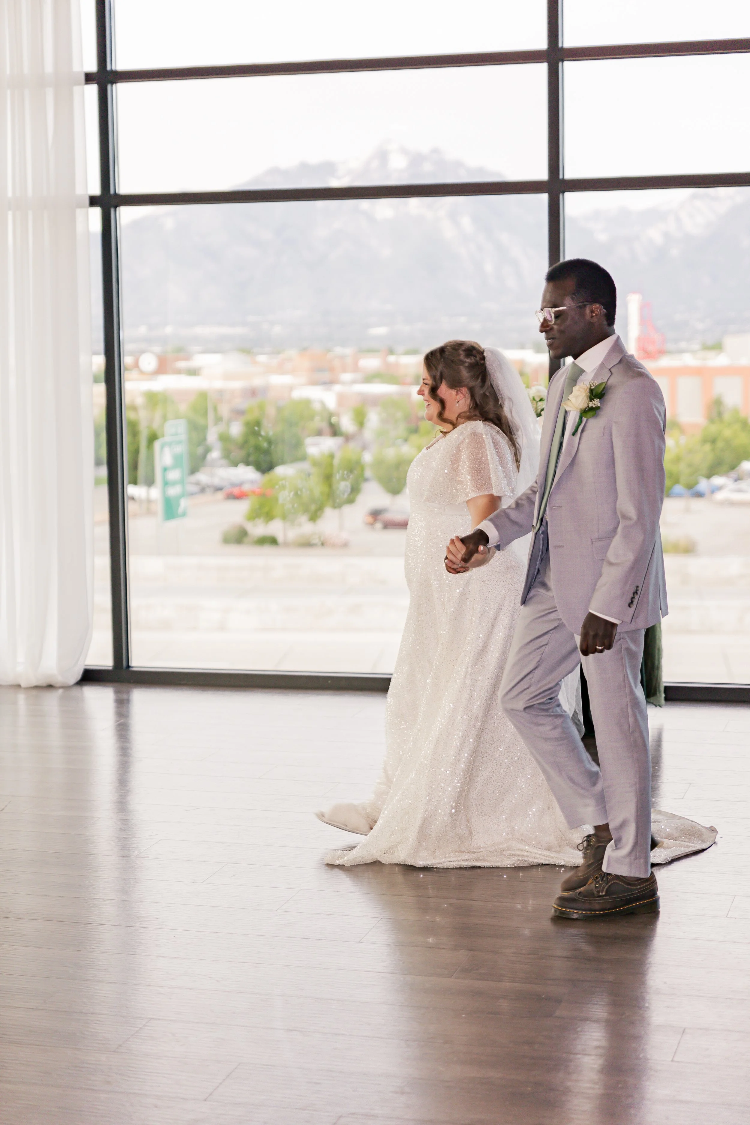 A bride and groom holding hands and walking inside a wedding venue with large windows and a mountain view.